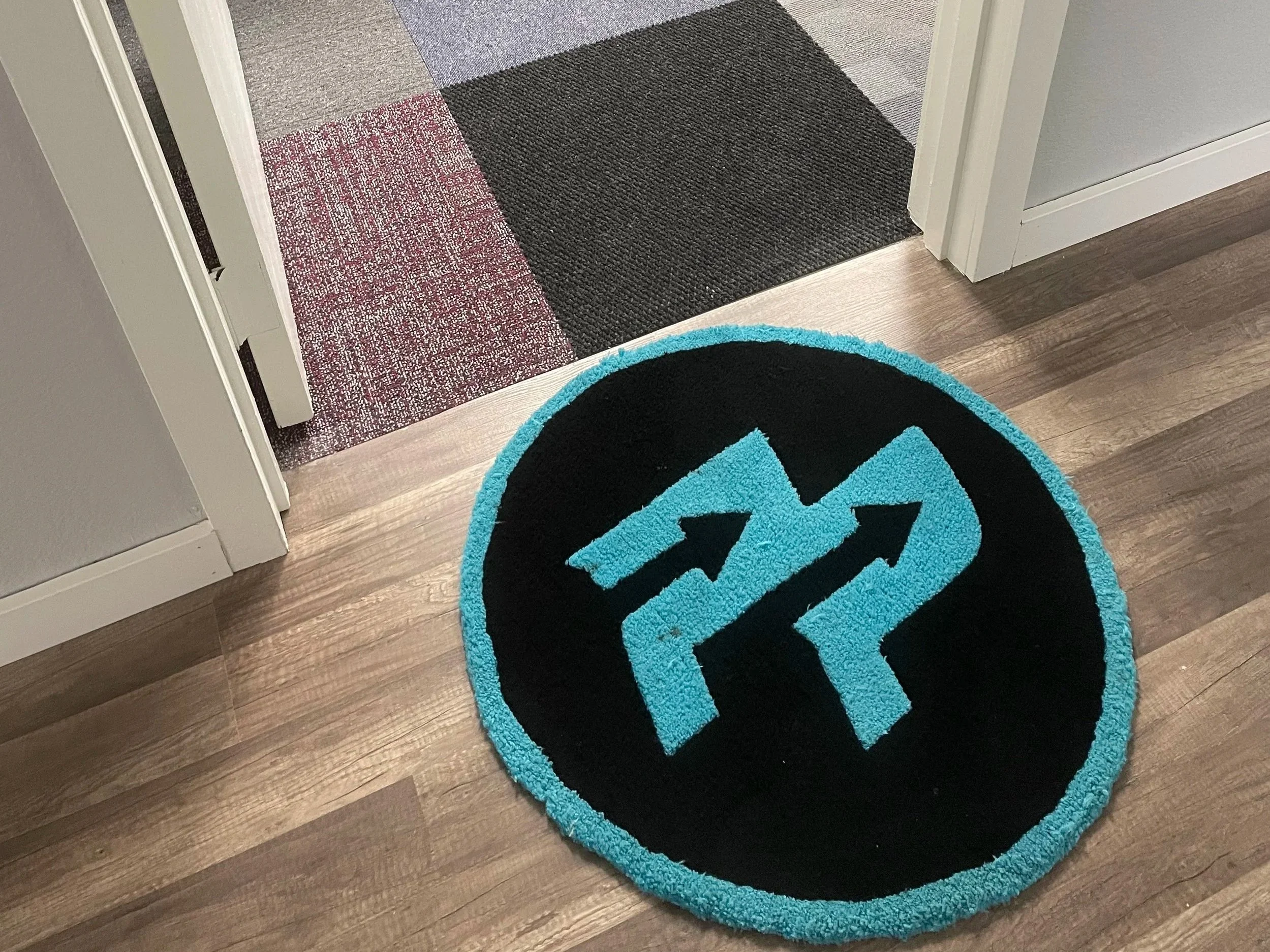 A round black and turquoise rug with a geometric design on a wooden floor near a doorway with carpeted threshold.