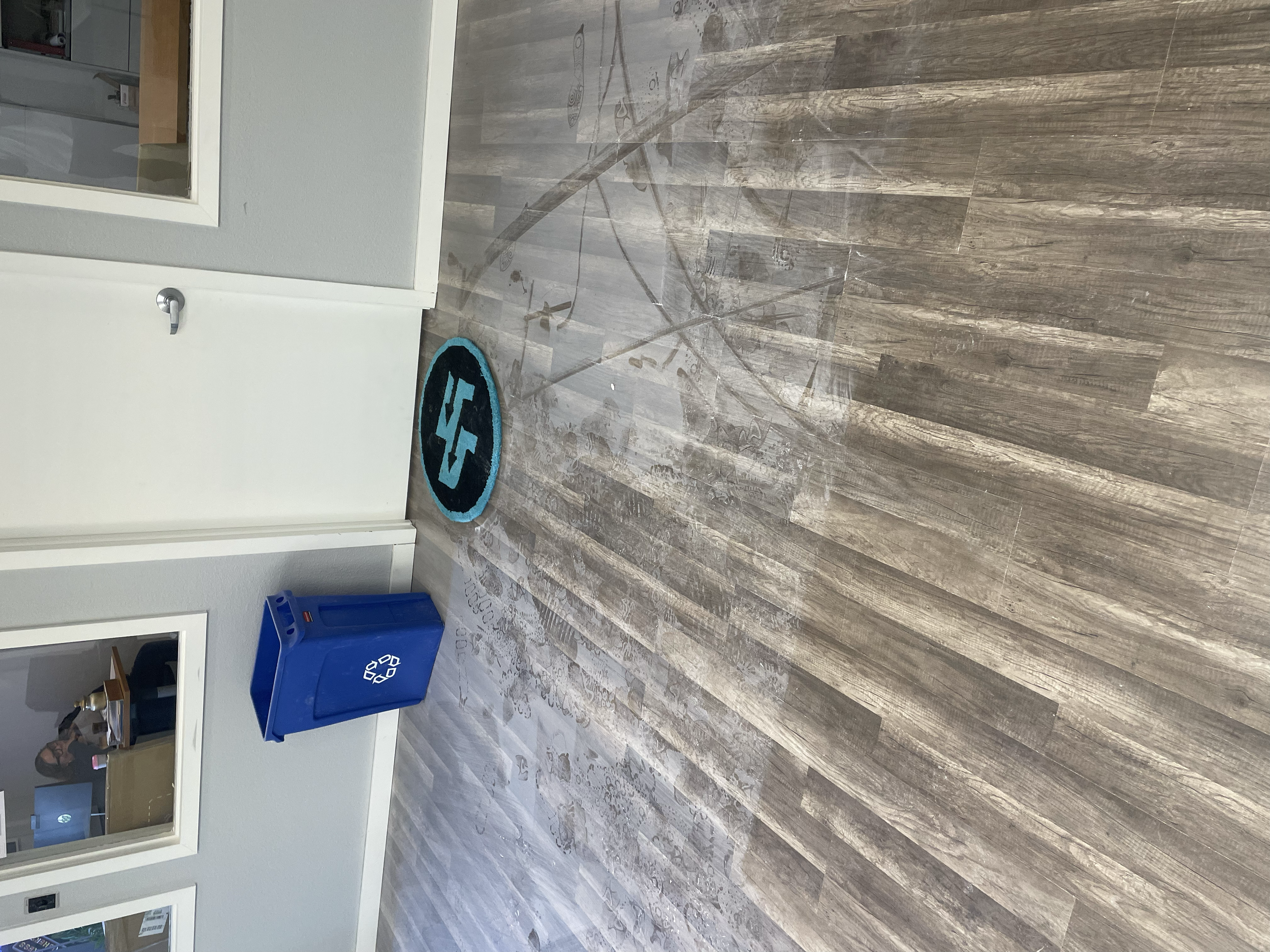 Wet floor with water pooled on wood laminate flooring, a blue recycling bin, and a black and blue rug with a lightning bolt symbol near white walls and a glass display case.