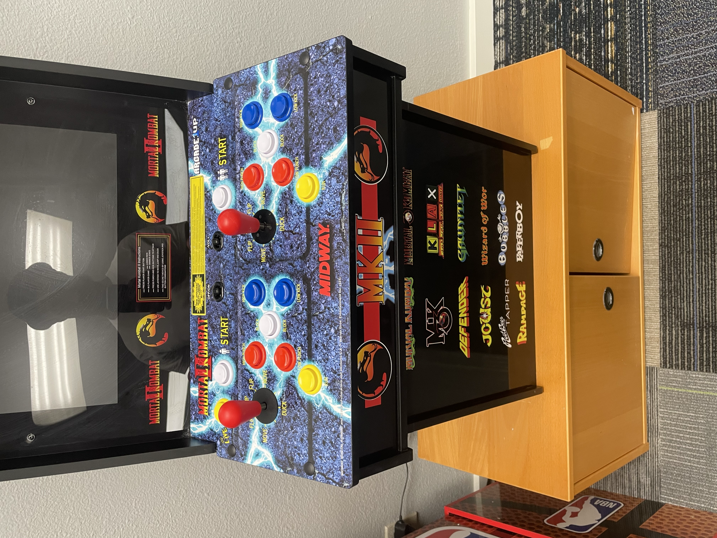 Up-close view of a Mortal Kombat arcade game with colorful controls, including buttons and joysticks, set on a wooden cabinet in an indoor space.