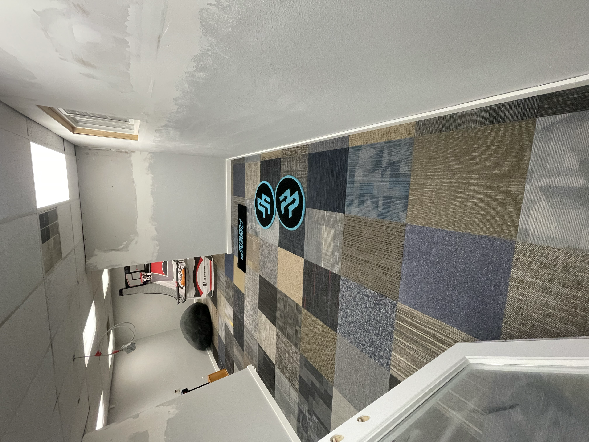 An indoor room with a patterned carpeted floor, partially painted white walls, and a suspended ceiling with fluorescent lights. There is a basketball hoop on one wall, a large black exercise ball, and some exercise equipment in the background. Two bl