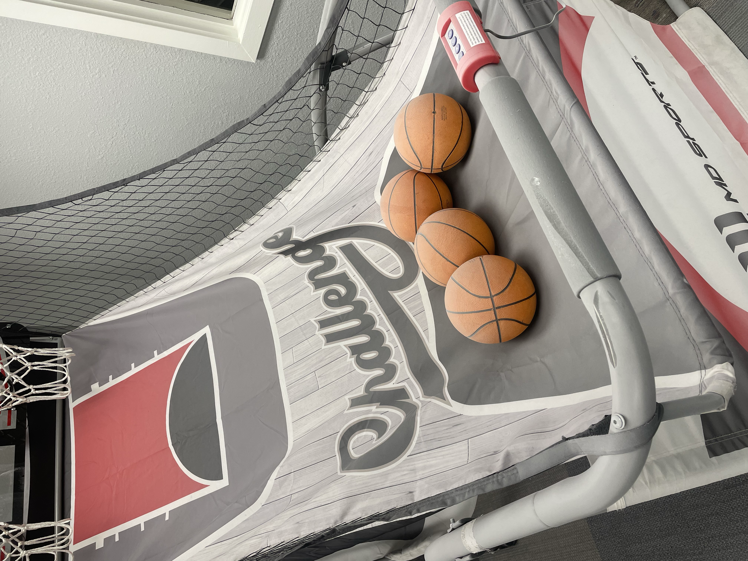 Four basketballs on a game backboard with a mini basketball hoop, a roller, and a plastic bag with the words 'Land Sport' on it, set up on a balcony or outdoor space against a wall with a window.