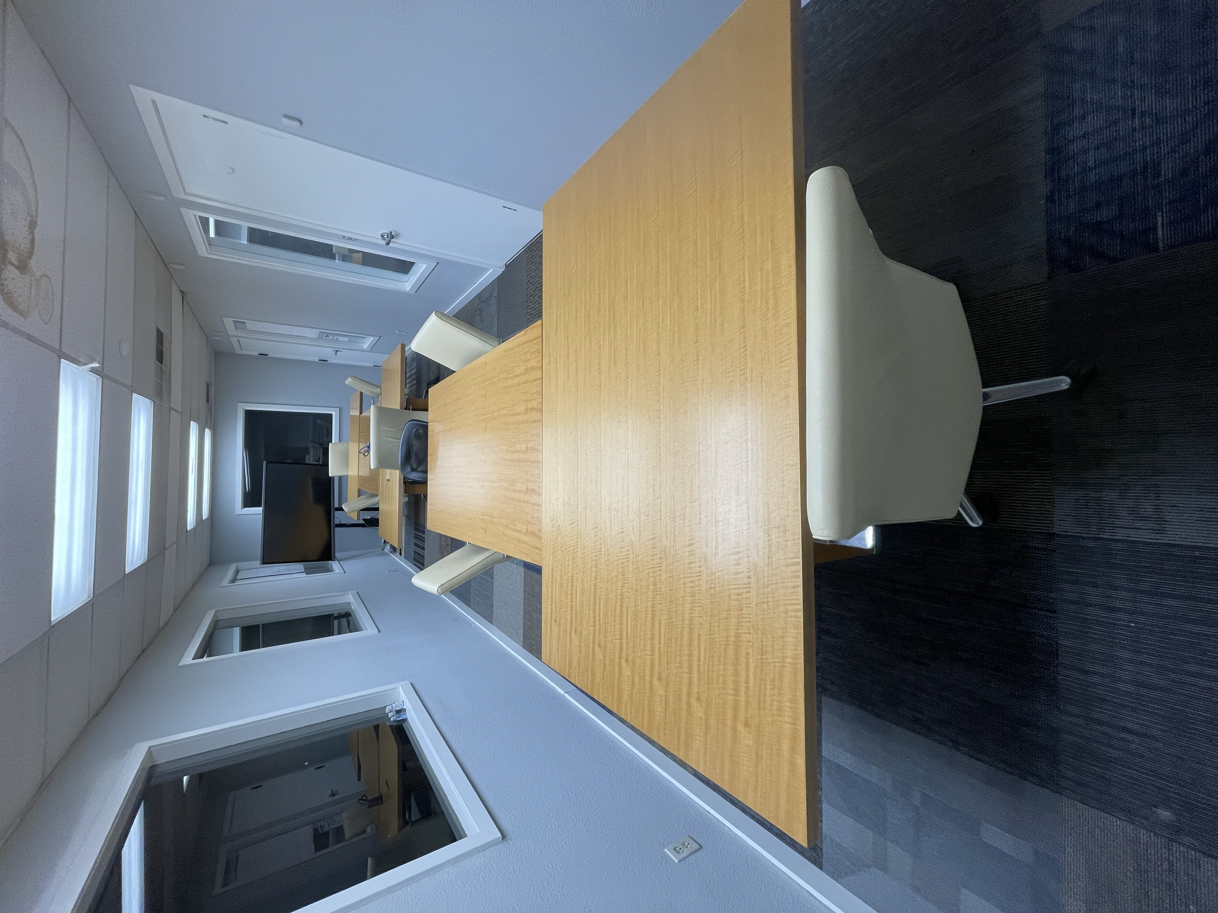 A conference room with a long wooden table and beige chairs, a large monitor, windows, and a door.