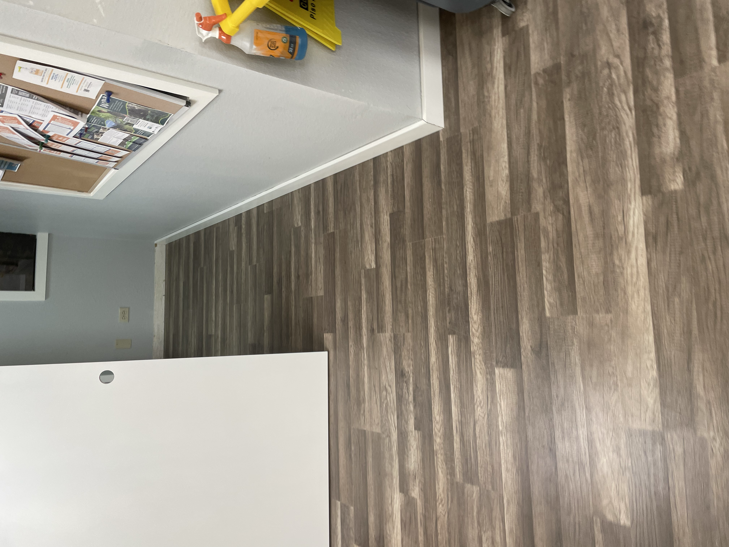 An indoor room with a white wall, wood flooring, and a white countertop. There are yellow cleaning supplies and an orange and white spray bottle on top of the counter. A bulletin board is mounted on the wall.