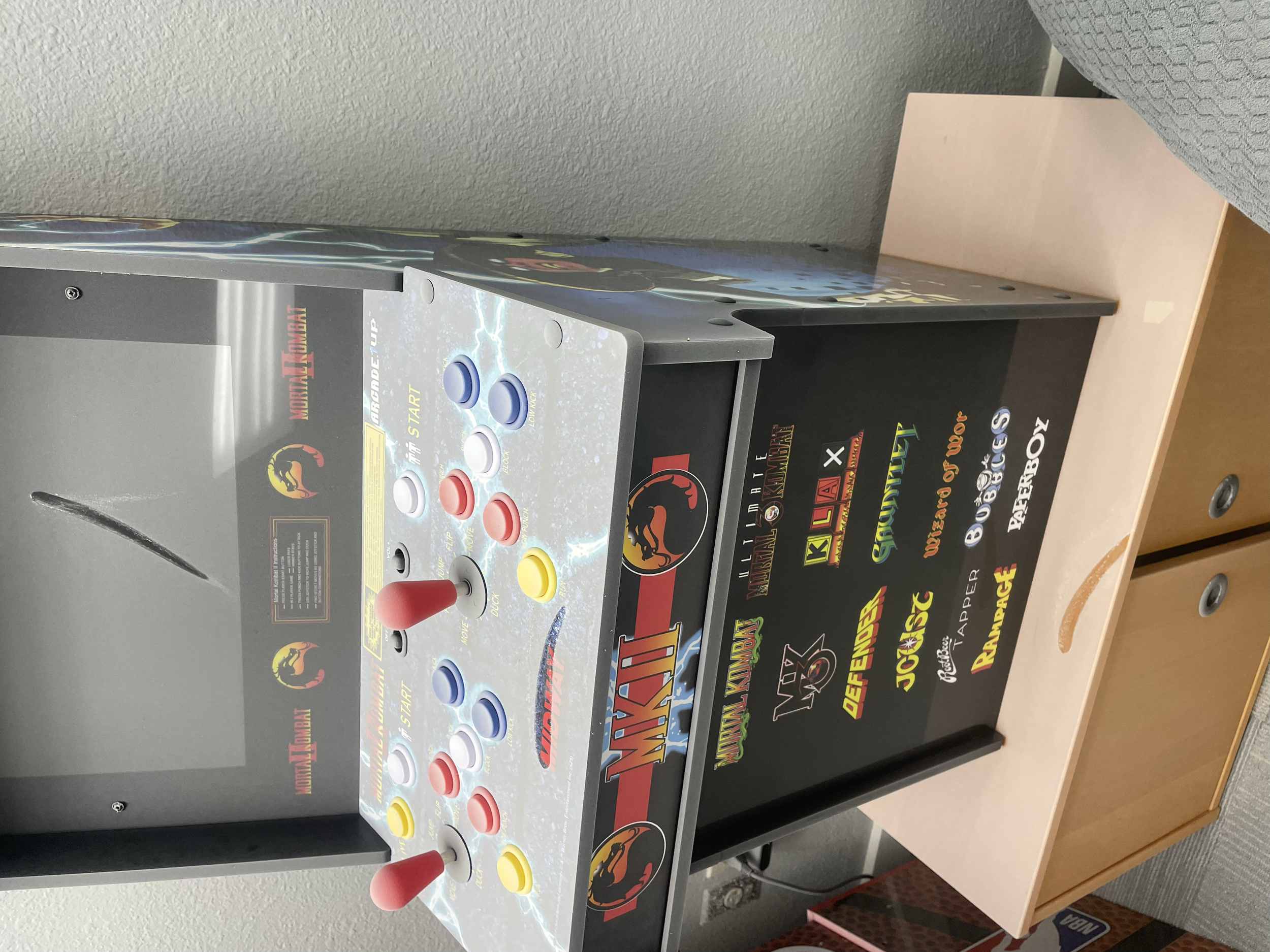 A Mortal Kombat arcade game machine with colorful buttons, joysticks, and a screen, placed on a wooden table.