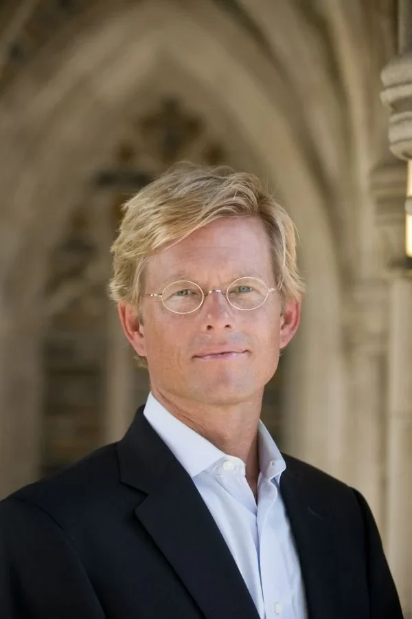 A man with blonde hair and glasses wearing a black suit jacket and white shirt standing in front of a stone archway.