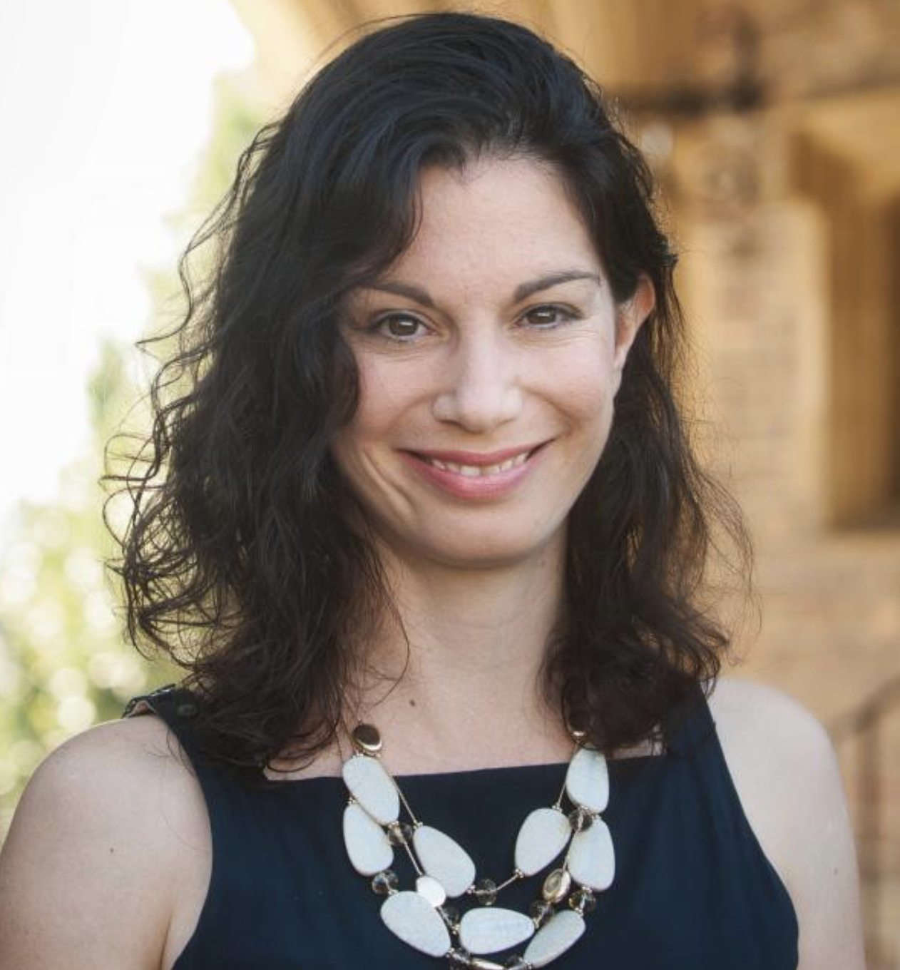 A woman with black, wavy hair smiling while wearing a black sleeveless top and a large white statement necklace.