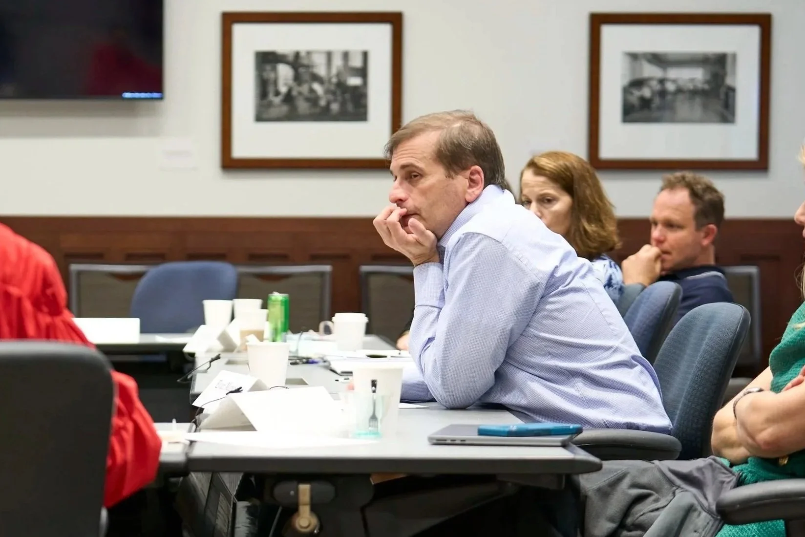A group of people engaged in a discussion during a meeting in a conference room. The man in the blue shirt is listening attentively.