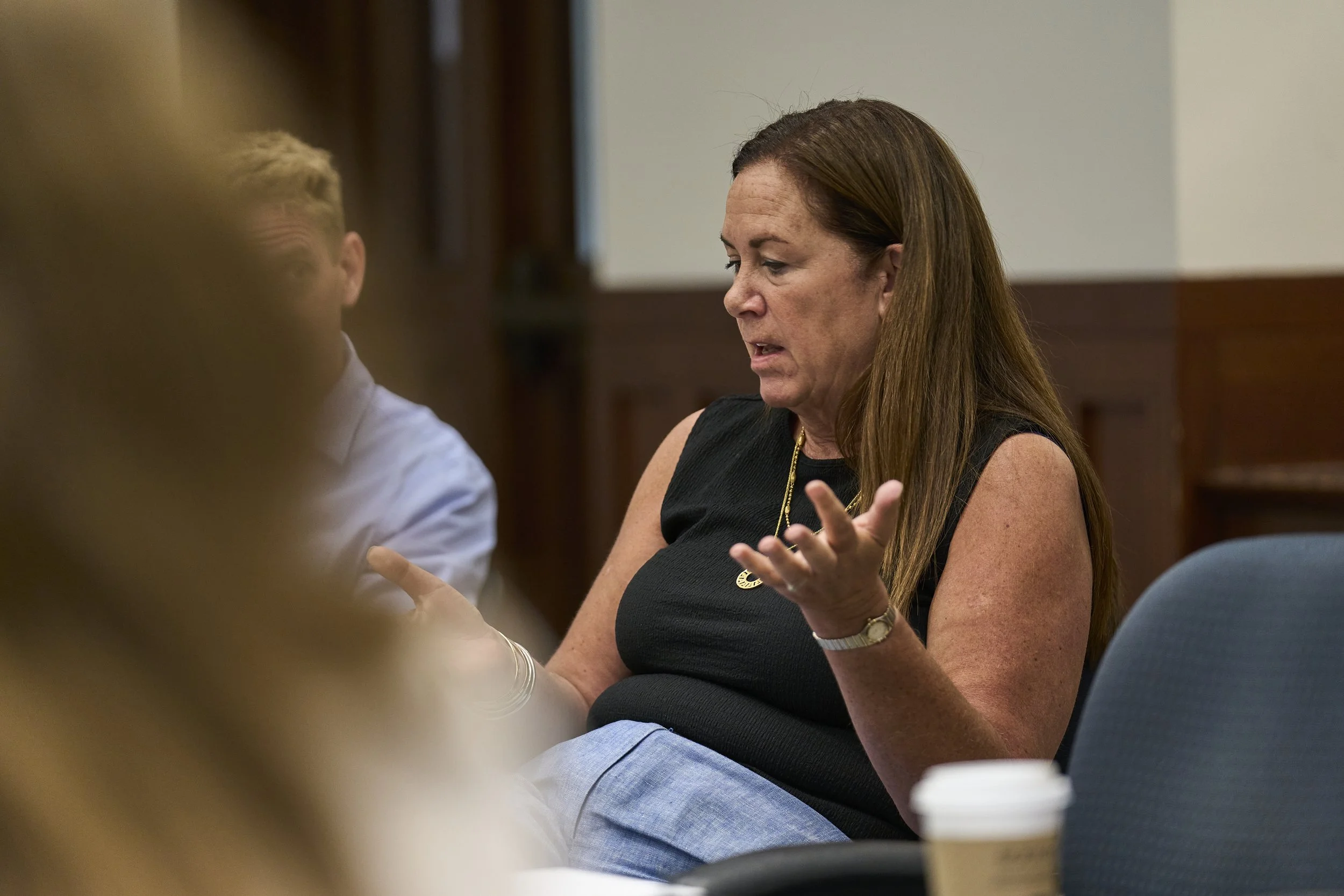 A woman wearing a black top gesturing with her hands while seated at a table during a discussion or meeting.