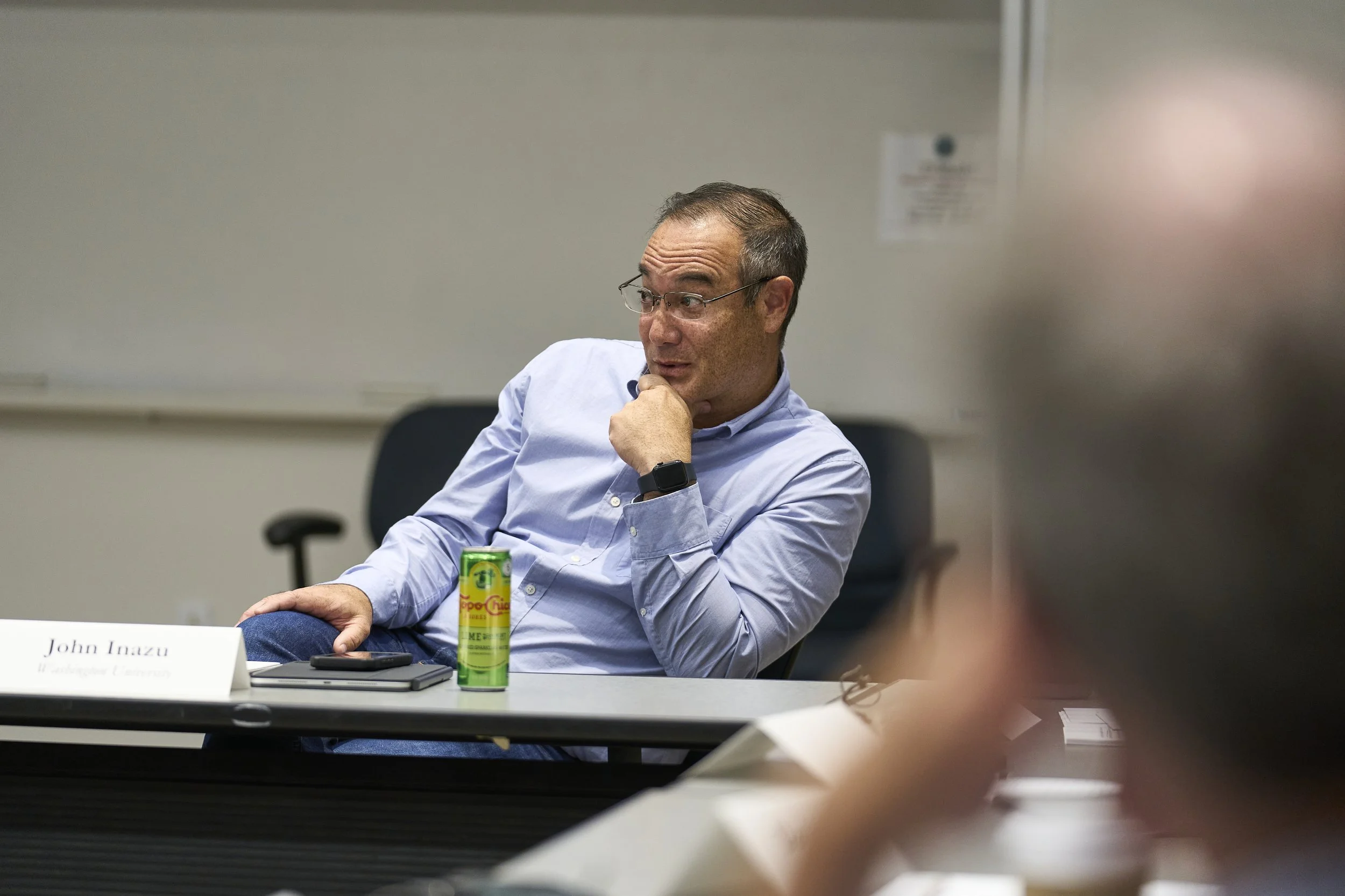 A man with glasses and a light blue button-up shirt sitting at a conference table, resting his chin on his hand, with a can of food, a phone, and a notebook in front of him.