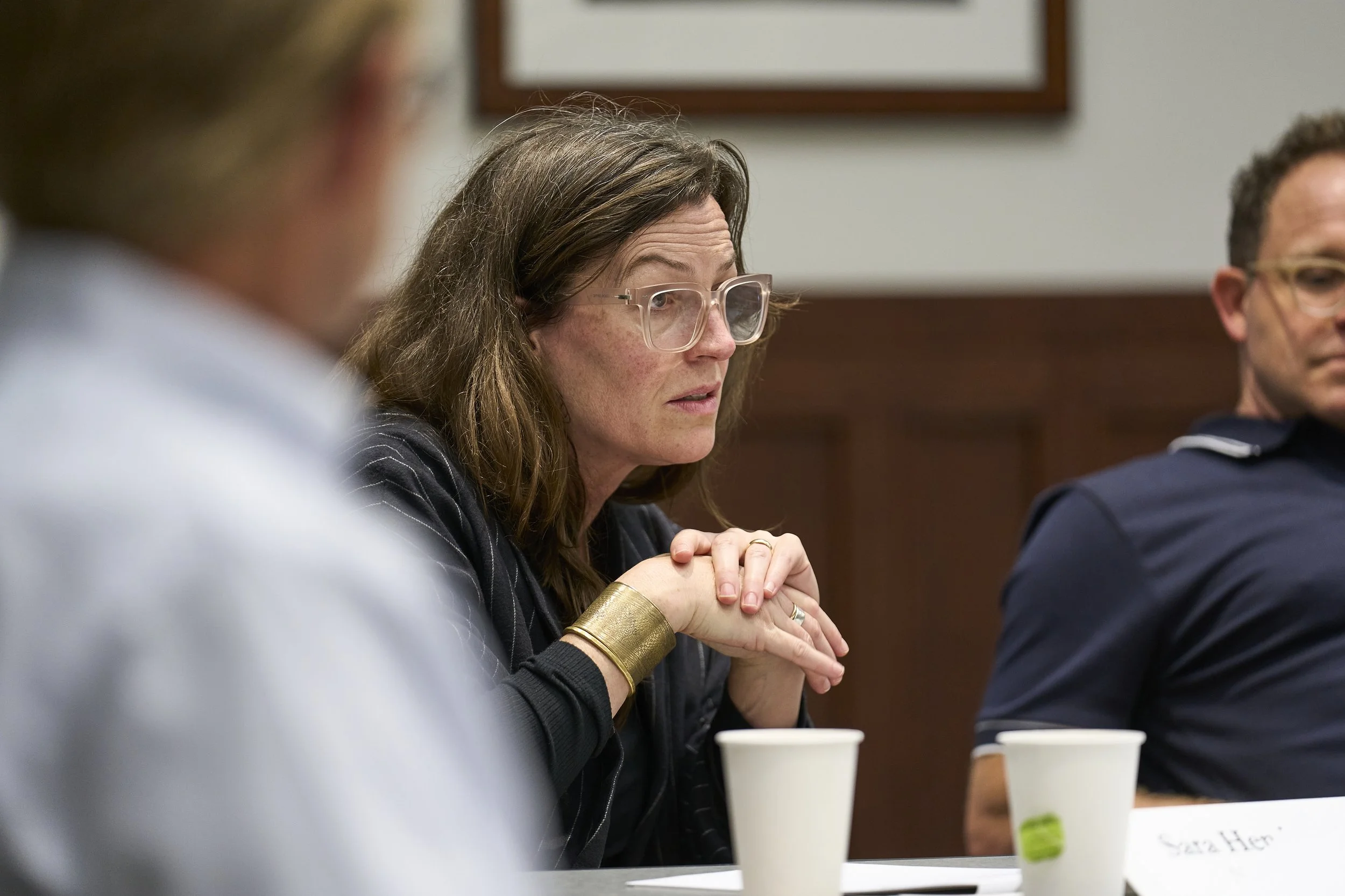 A woman with glasses in black blazer with thin white stripes, hands folded, leaning into to the discussion listening attentively.