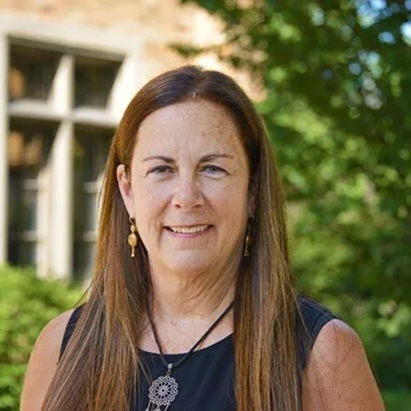 A woman with long brown hair and light skin, wearing a black sleeveless top and gold earrings, smiling outdoors with green trees and a building in the background.