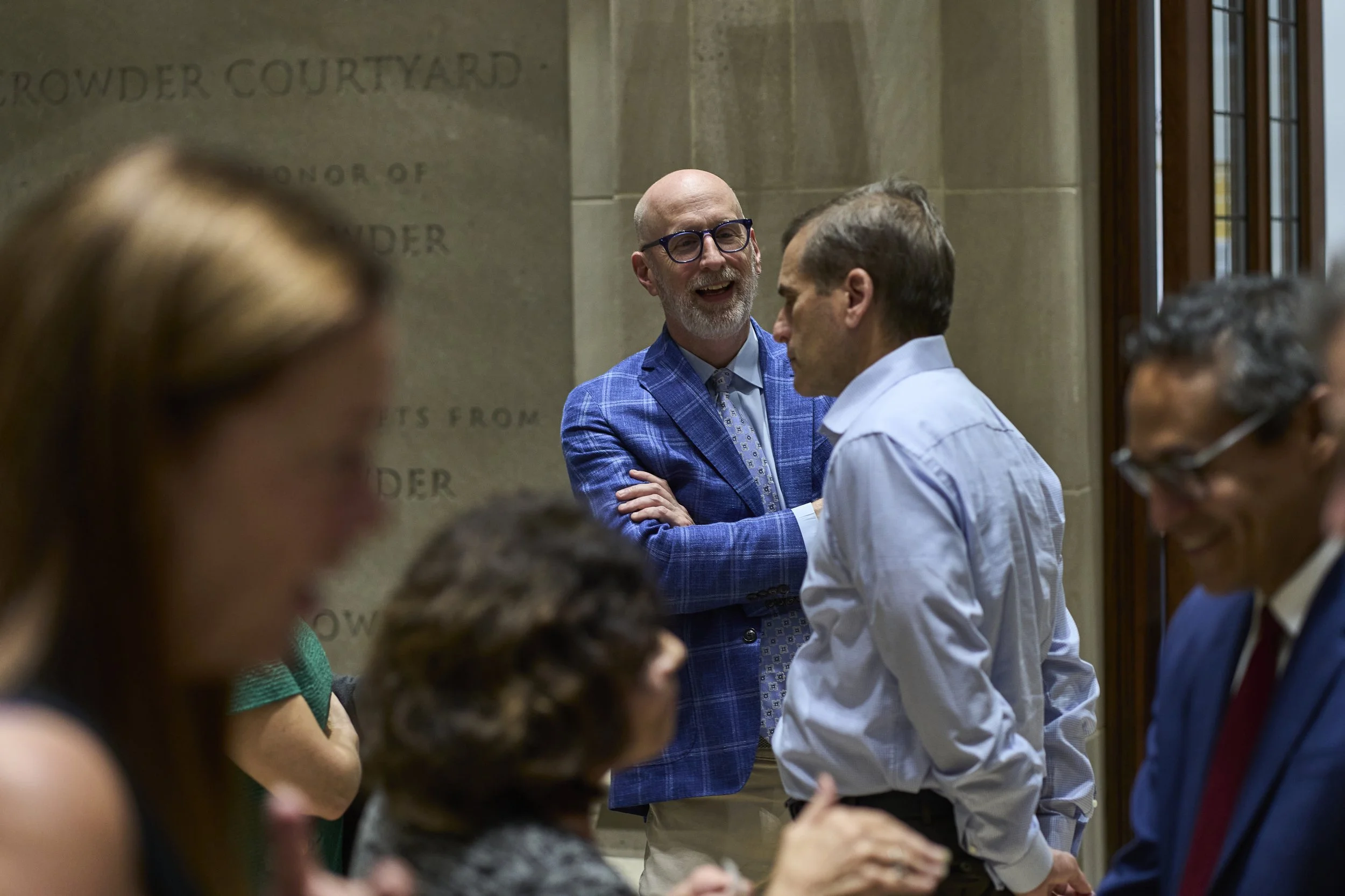A smiling man with glasses and a blue plaid blazer talking to a man in a white shirt, surrounded by other people engaged in conversation in an indoor setting.