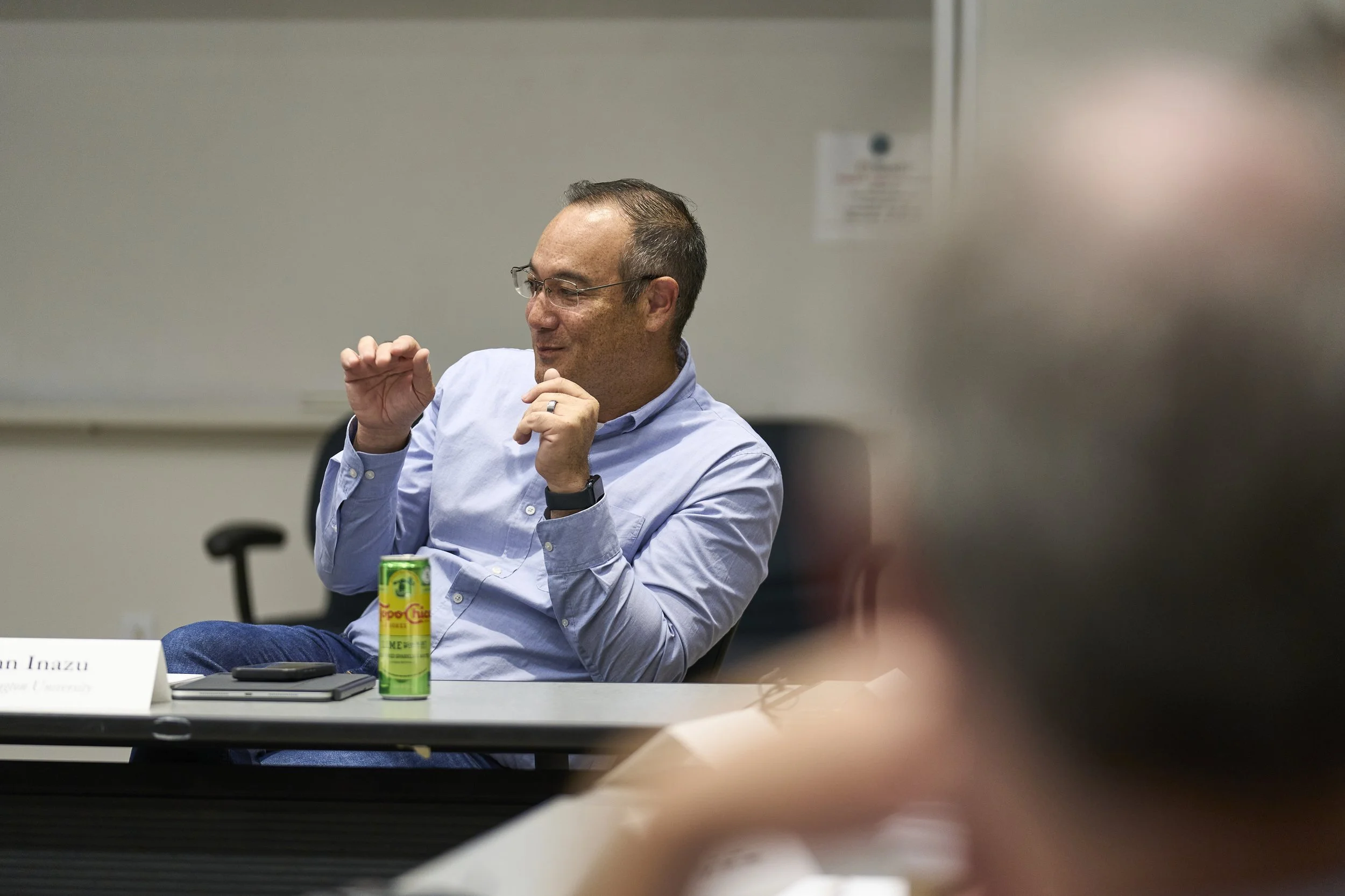 A man in a blue shirt smiling and gesturing with his hands during a discussion seated at the conference table during a meeting.