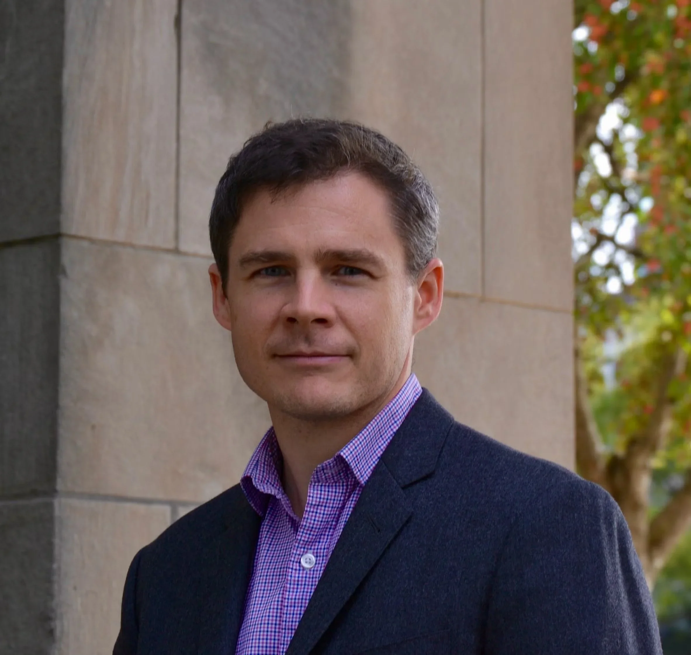A young man with short dark hair and light skin, wearing a dark blazer and a purple checkered shirt, standing outdoors near a stone building with greenery in the background.