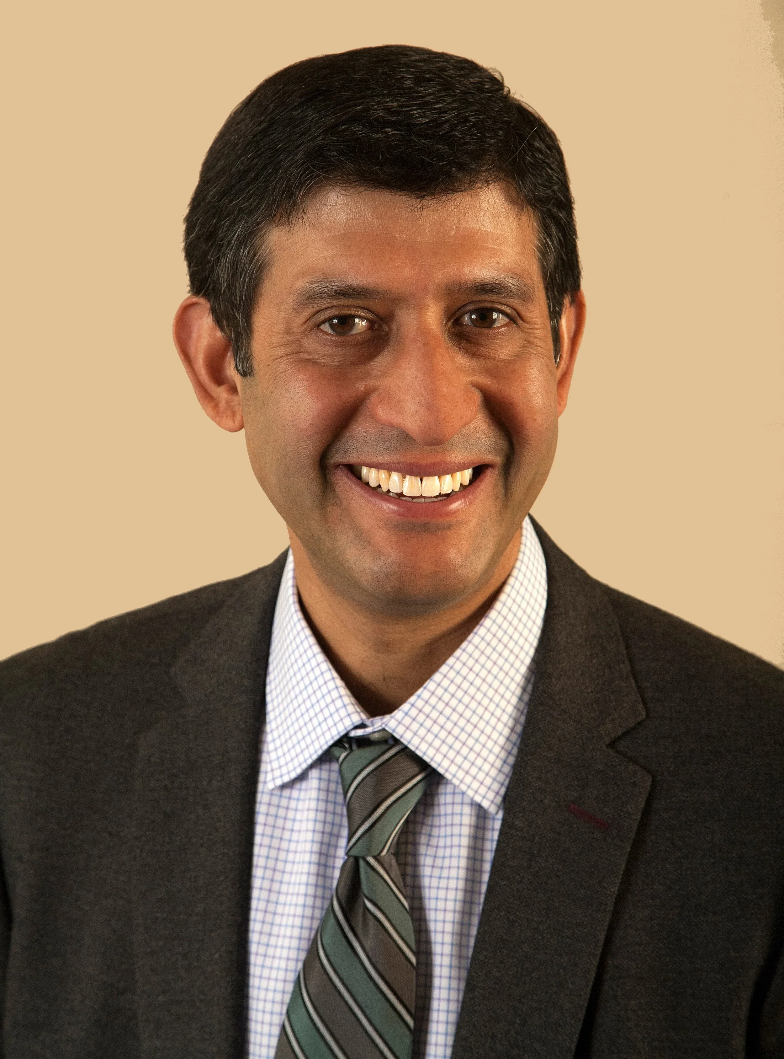 A smiling man wearing a dark suit, light checked shirt, and striped tie, against a plain beige background.