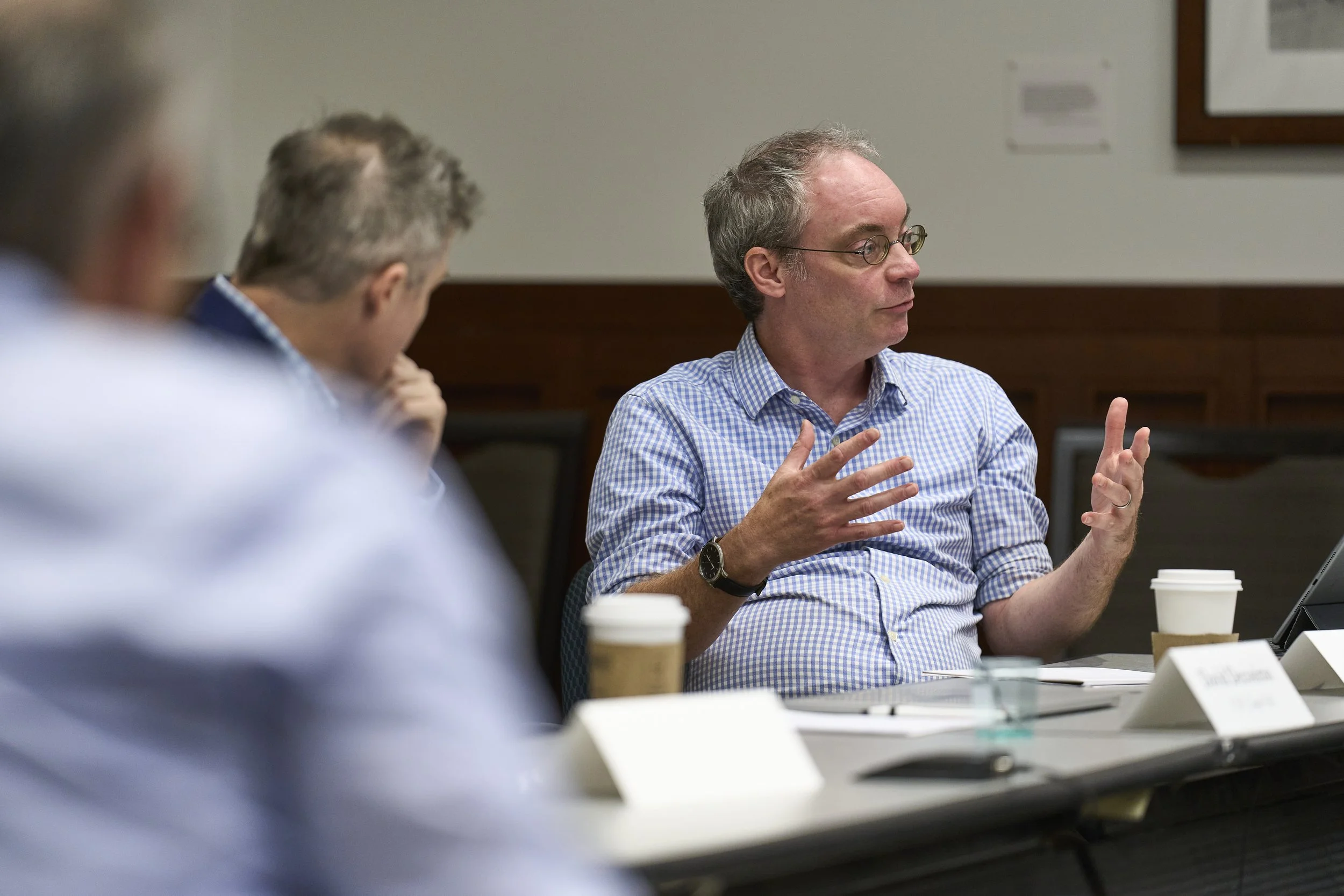 A man in a blue checkered shirt is speaking at a meeting, gesturing with his hands, with coffee cups and documents on the table. Two other people are partially visible in the foreground.