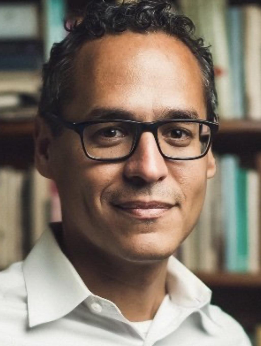 A man wearing glasses smiling in front of a bookshelf.