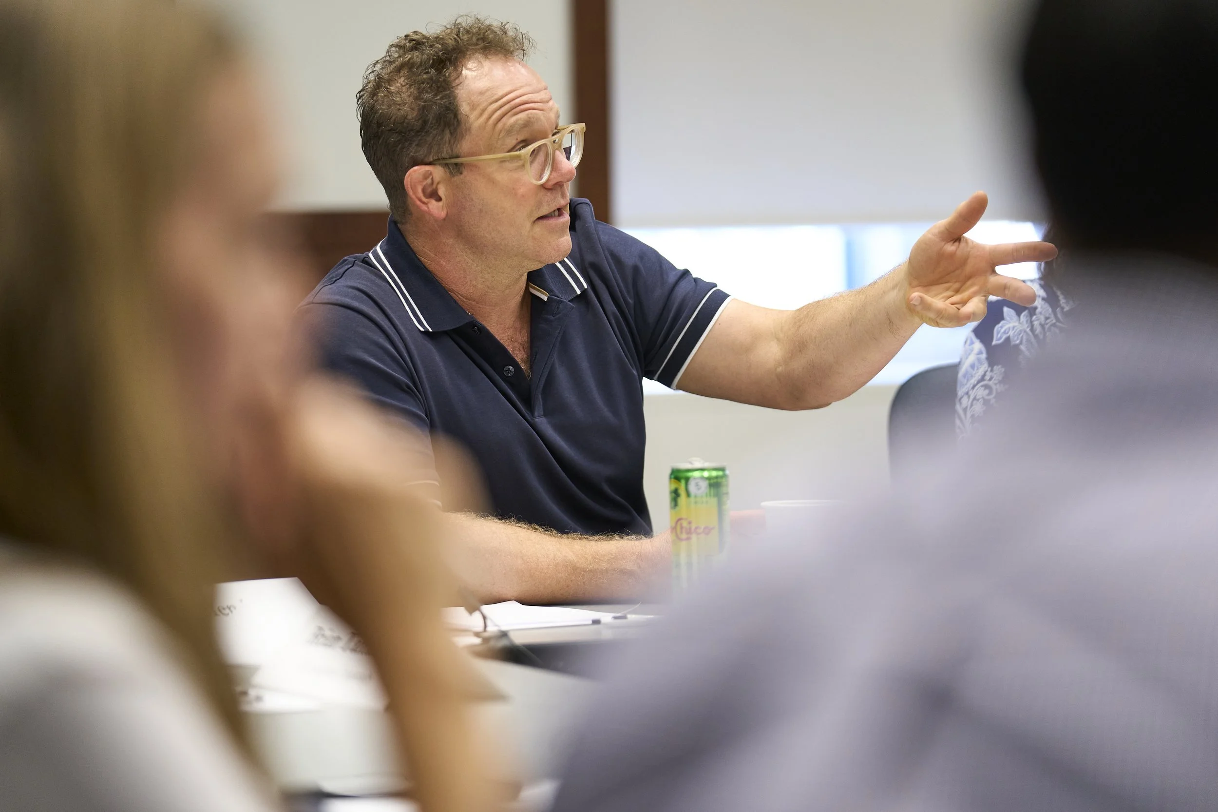 A man with glasses in a blue polo shirt speaking and gesturing with his hand during a meeting.