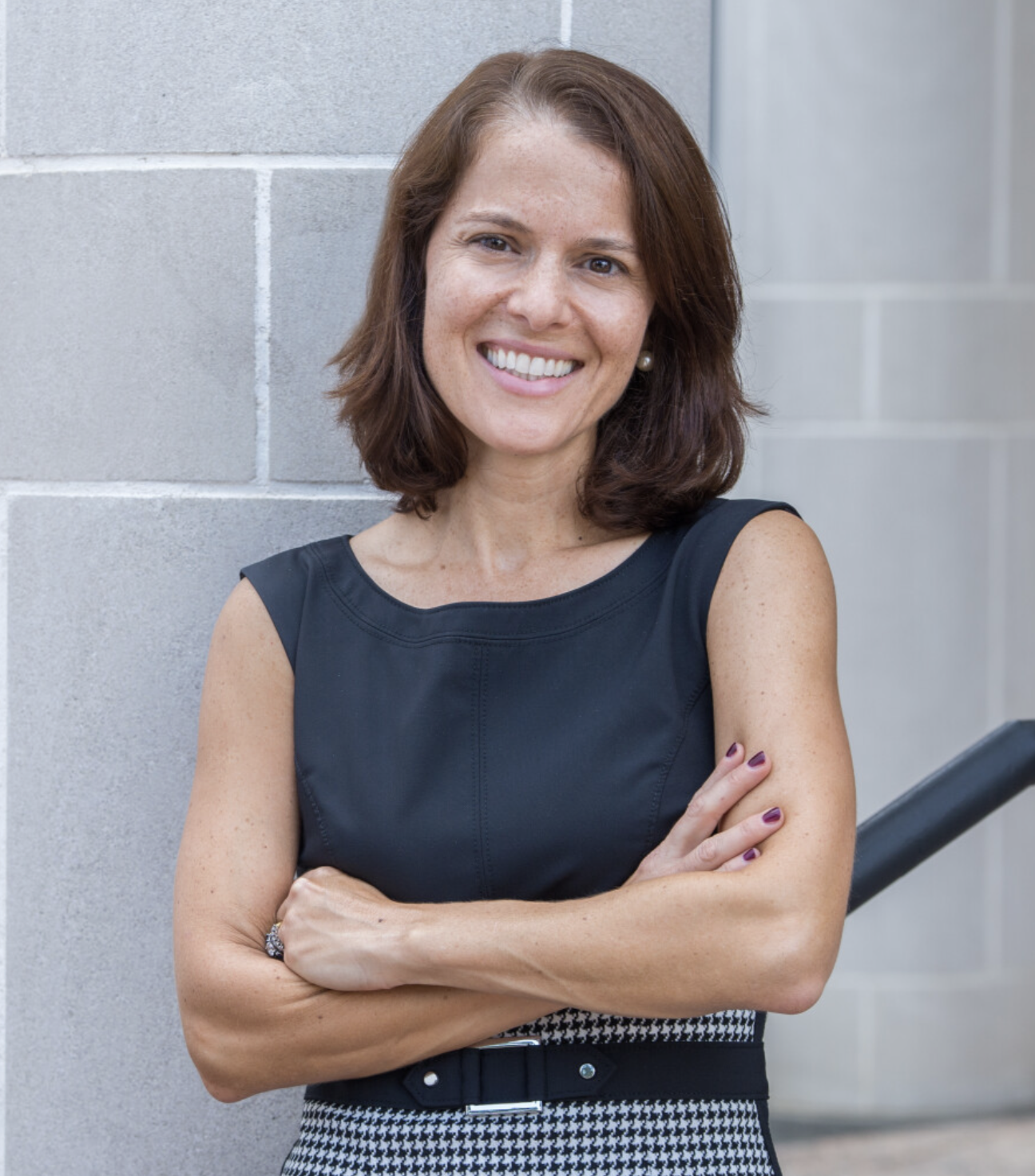 A smiling woman with brown hair, wearing a sleeveless black top and houndstooth skirt, standing against a concrete wall with her arms crossed.
