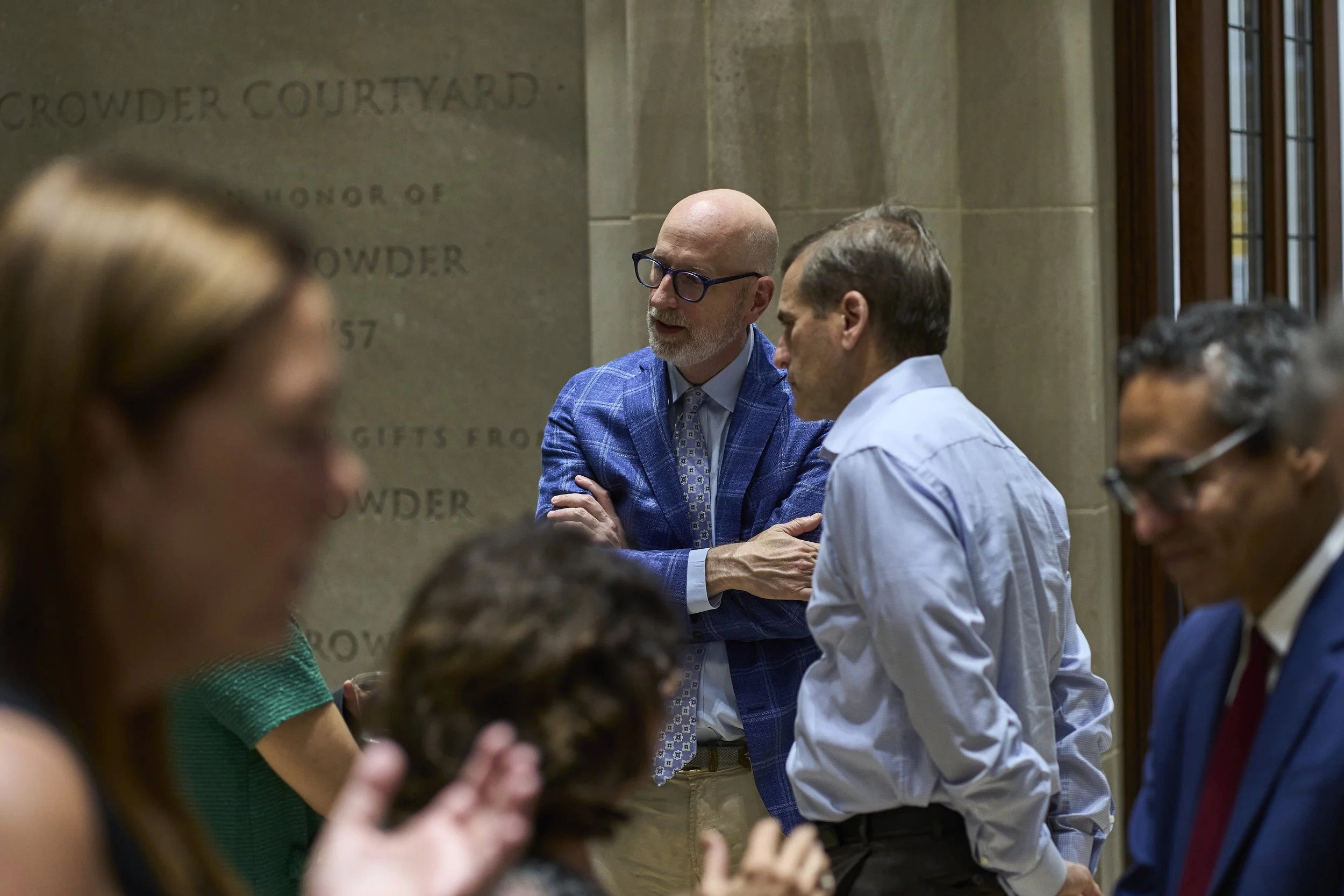 A group of five diverse people engaged in conversation in an indoor setting, with a stone wall behind them that has some engraved text.