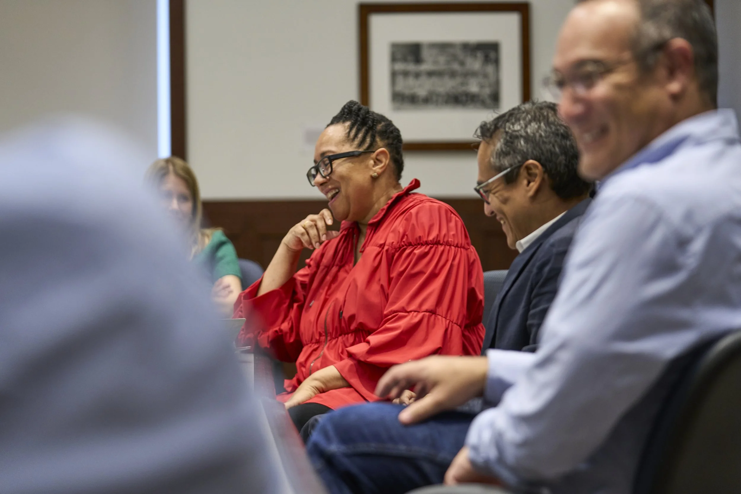 A woman wearing a red jacket and glasses laughing with her hand on her chin as colleagues around her are also smiling at each other.