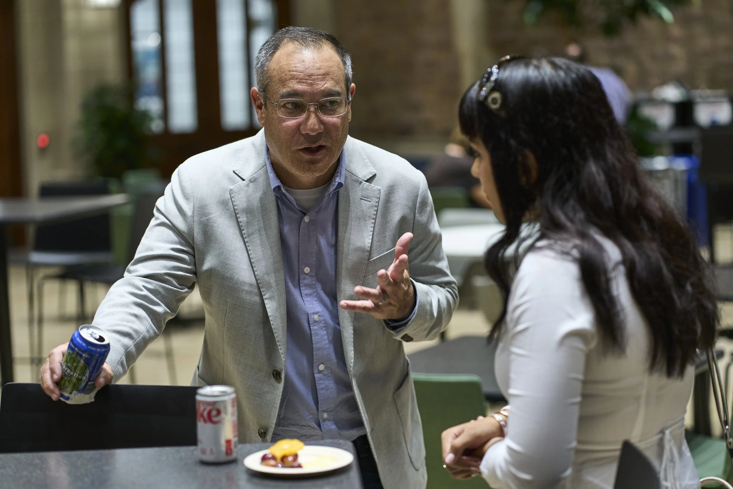 Two people having a conversation in a casual indoor setting with tables, chairs, and brick walls. One person, wearing a light gray blazer and glasses, is gesturing with his hand and holding a soda can in his other hand. The other person, with long da