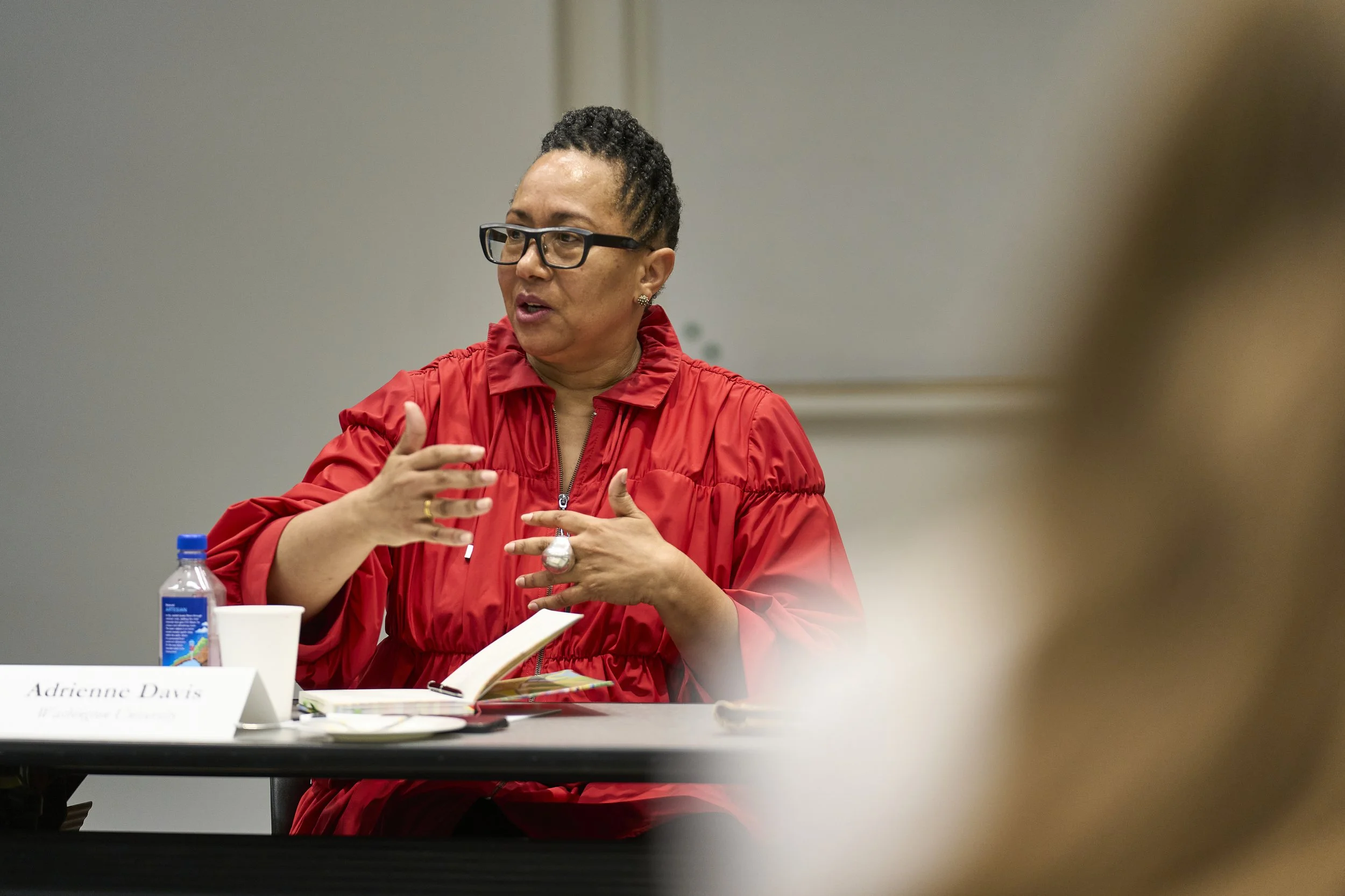 A woman wearing a red blouse and glasses, speaking and gesturing with her hands while seated at a table during a discussion.