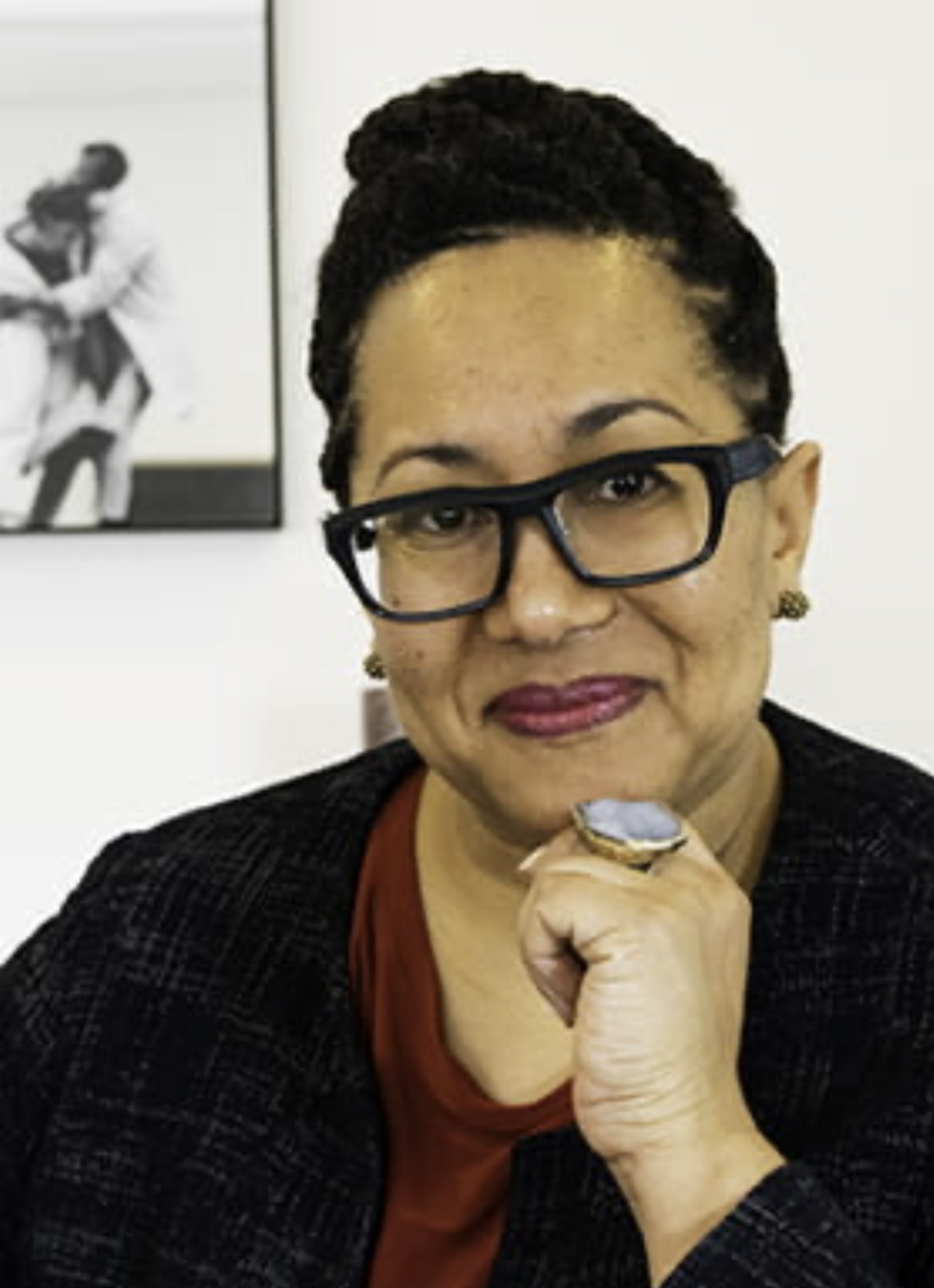 A woman with glasses, wearing a black blazer and red top, sitting indoors with a black and white photo of two children in the background.