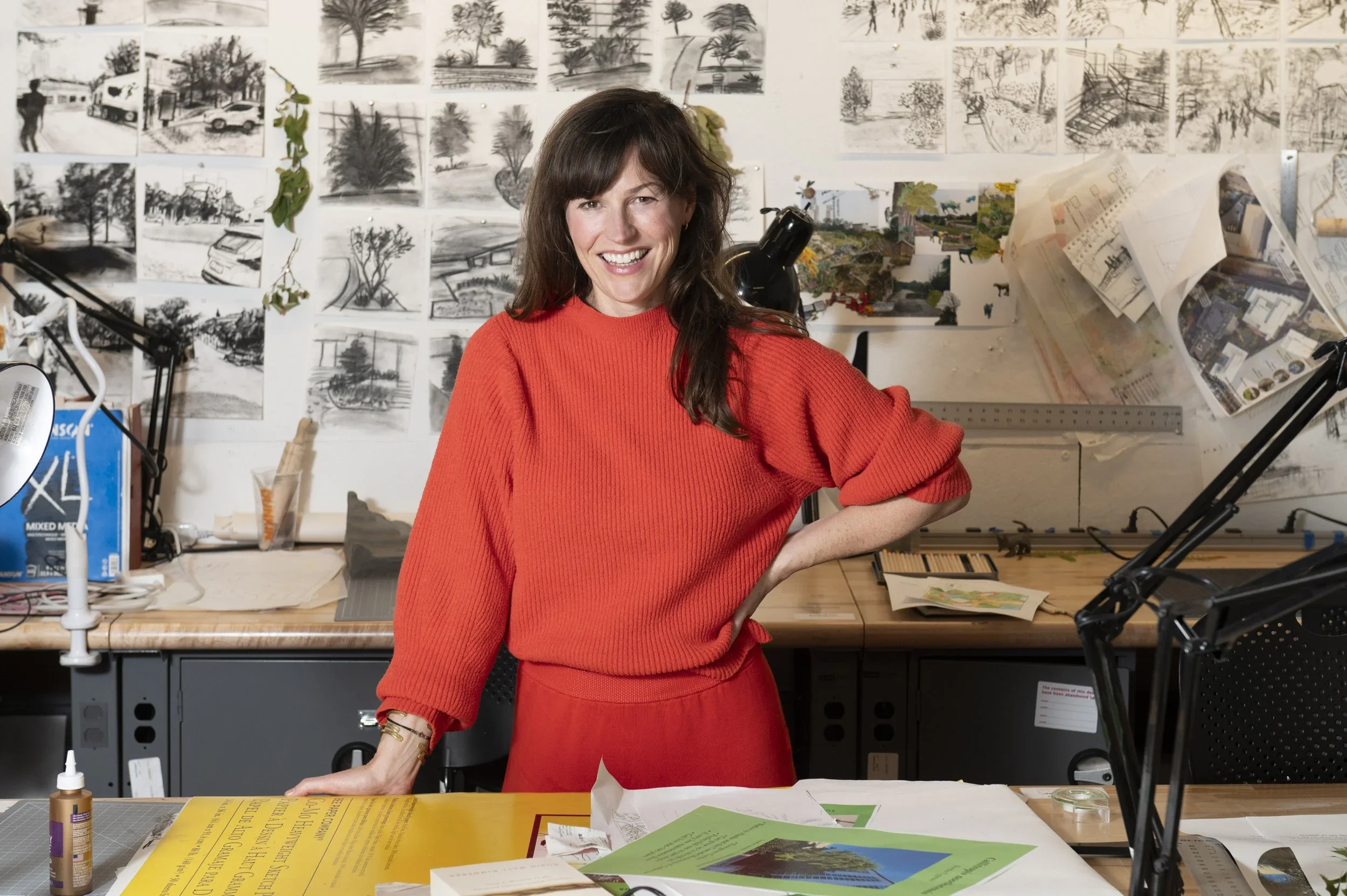 A woman in a red sweater standing in an art studio with drawings and sketches of trees and landscapes pinned to the wall behind her.