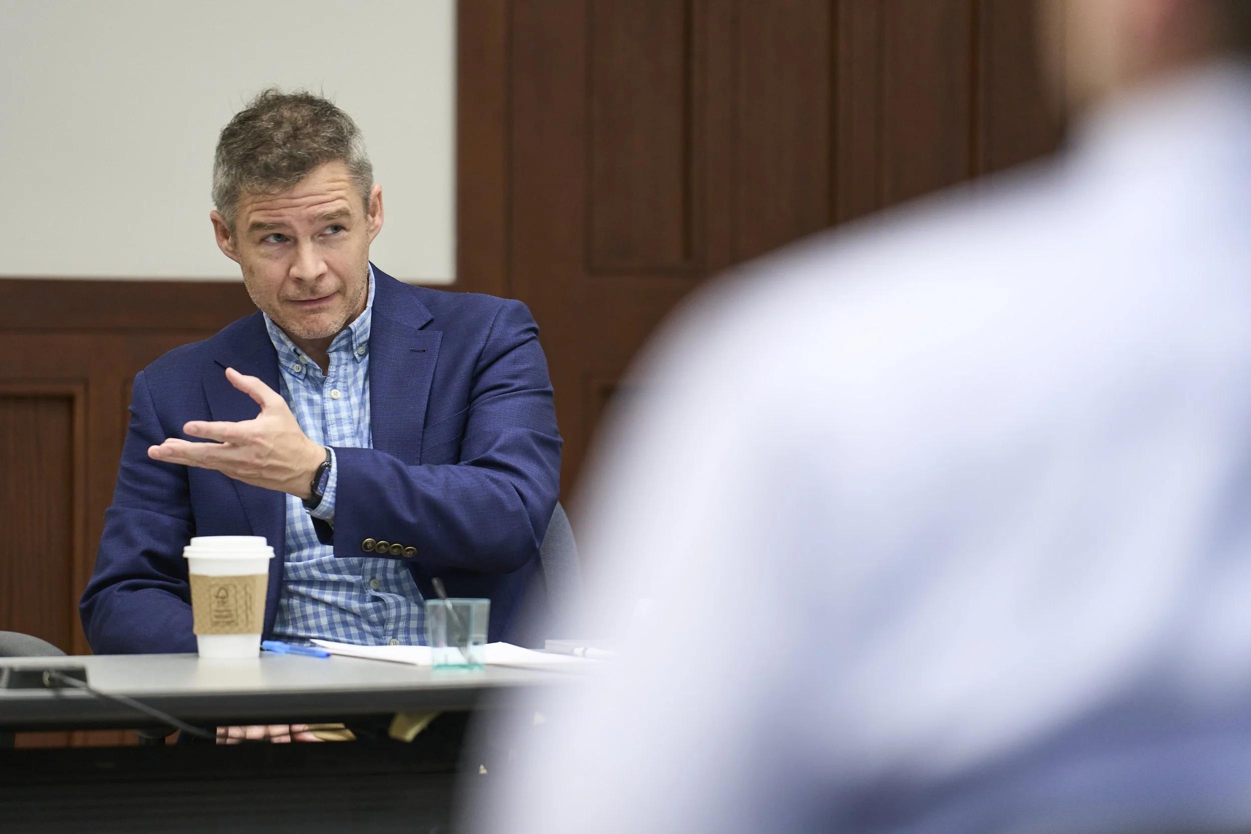 A man in a blue blazer and checked shirt gesturing with his hand in a discussion, sitting at a table with a coffee cup and papers, in a meeting room.