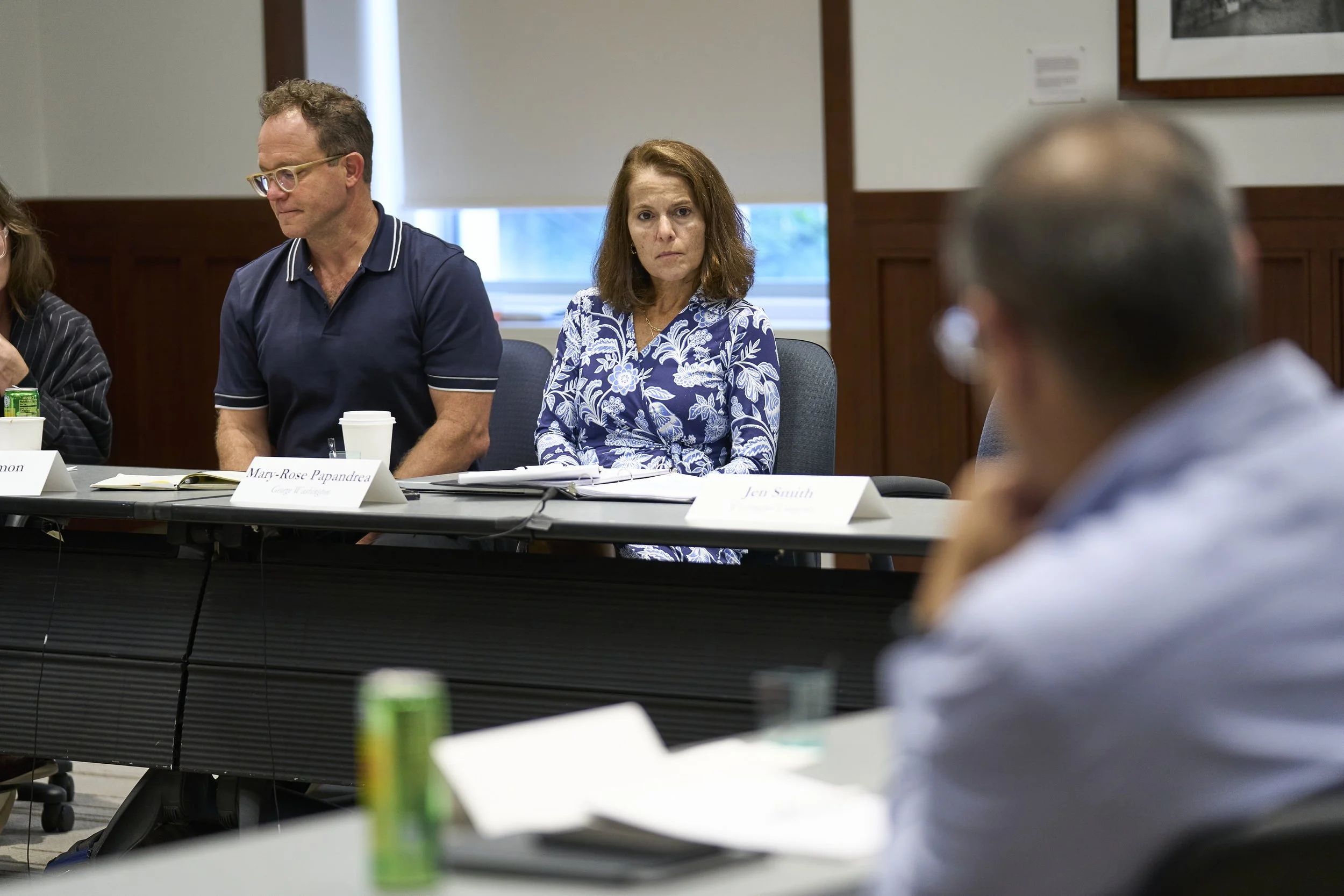 People attending a formal meeting or discussion in a conference room, sitting at a table with nameplates, notebooks, and drinks.