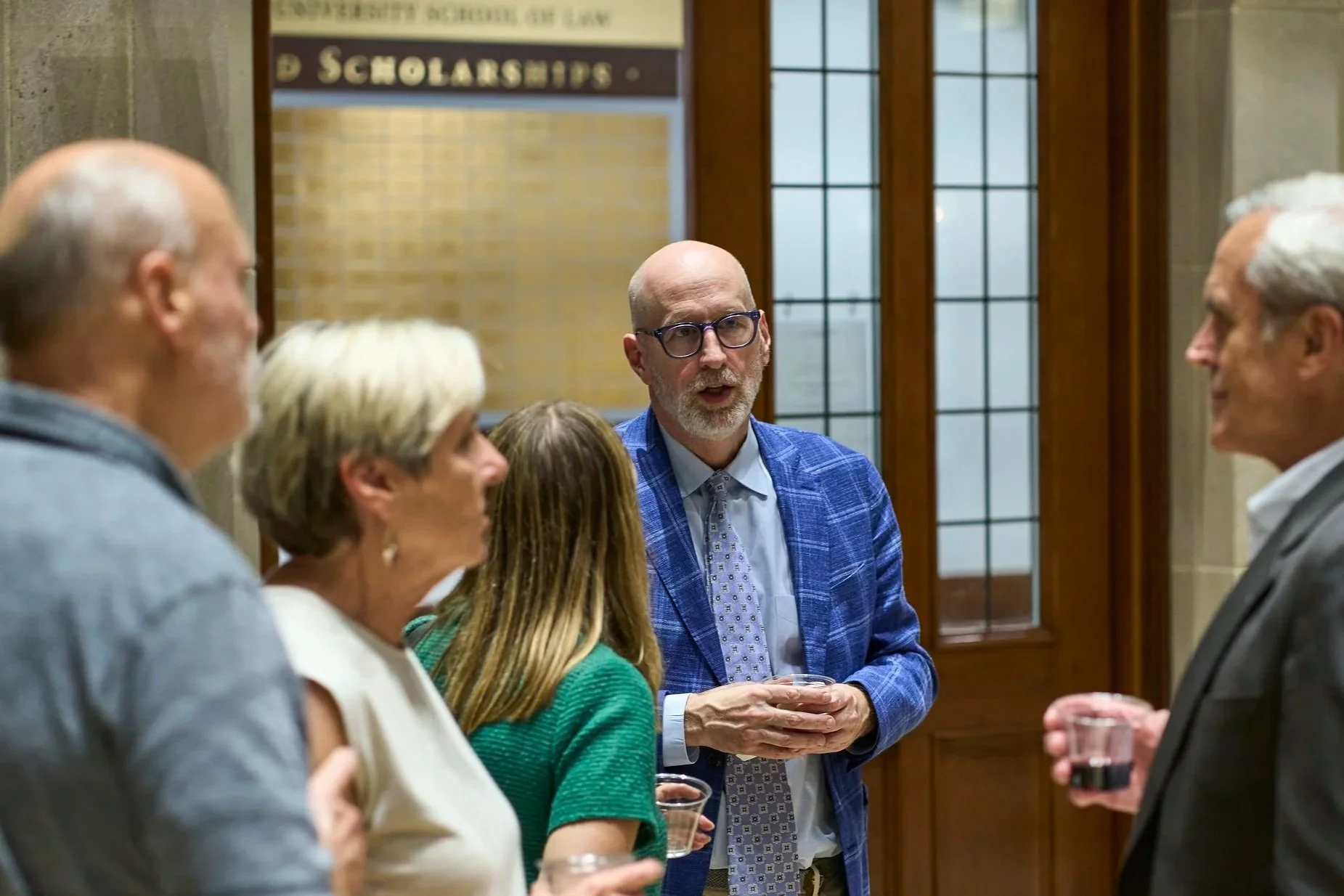 People in a conversation at a social event inside a building with large windows and wooden doors. The man in the center is wearing glasses and a blue suit jacket, holding a glass, and appears to be listening or speaking. The other four individuals are partially visible, engaged in the discussion.
