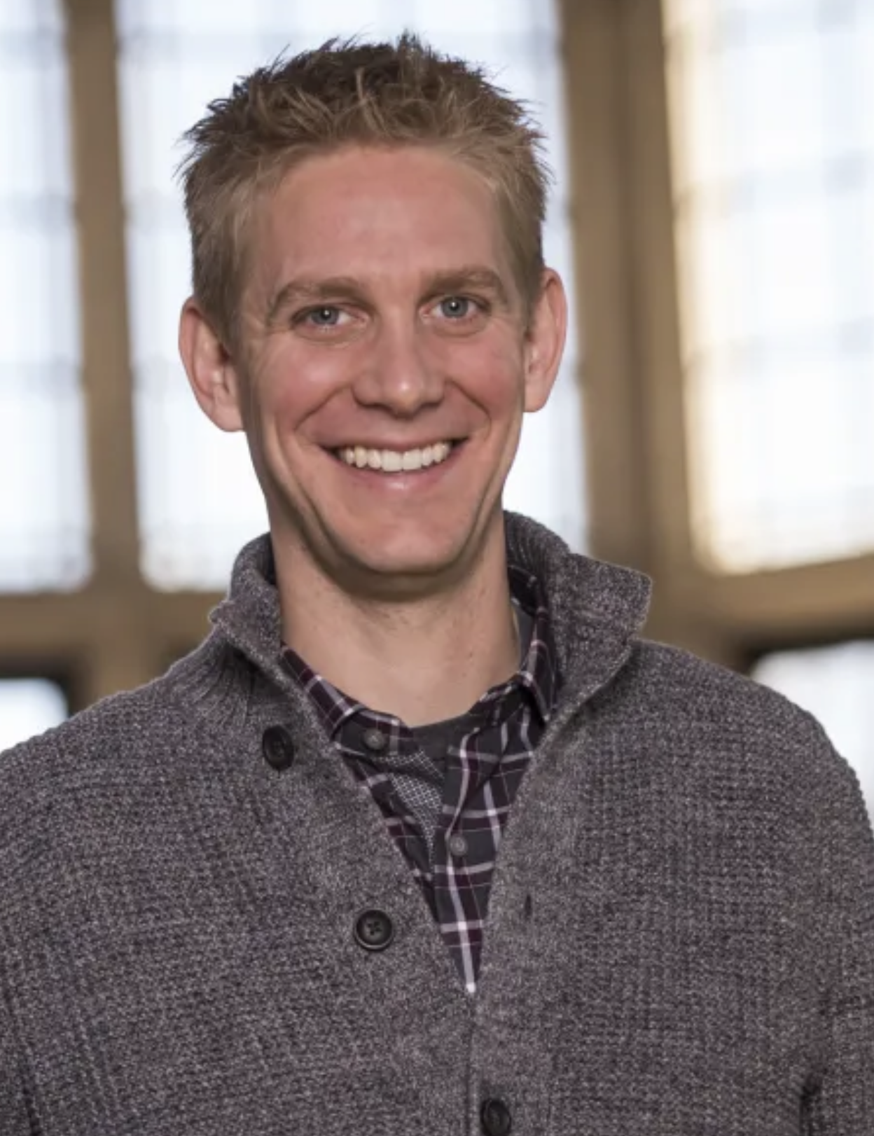 A smiling man with short, light brown hair, wearing a gray knitted cardigan over a plaid shirt, standing in front of a window with curtains.