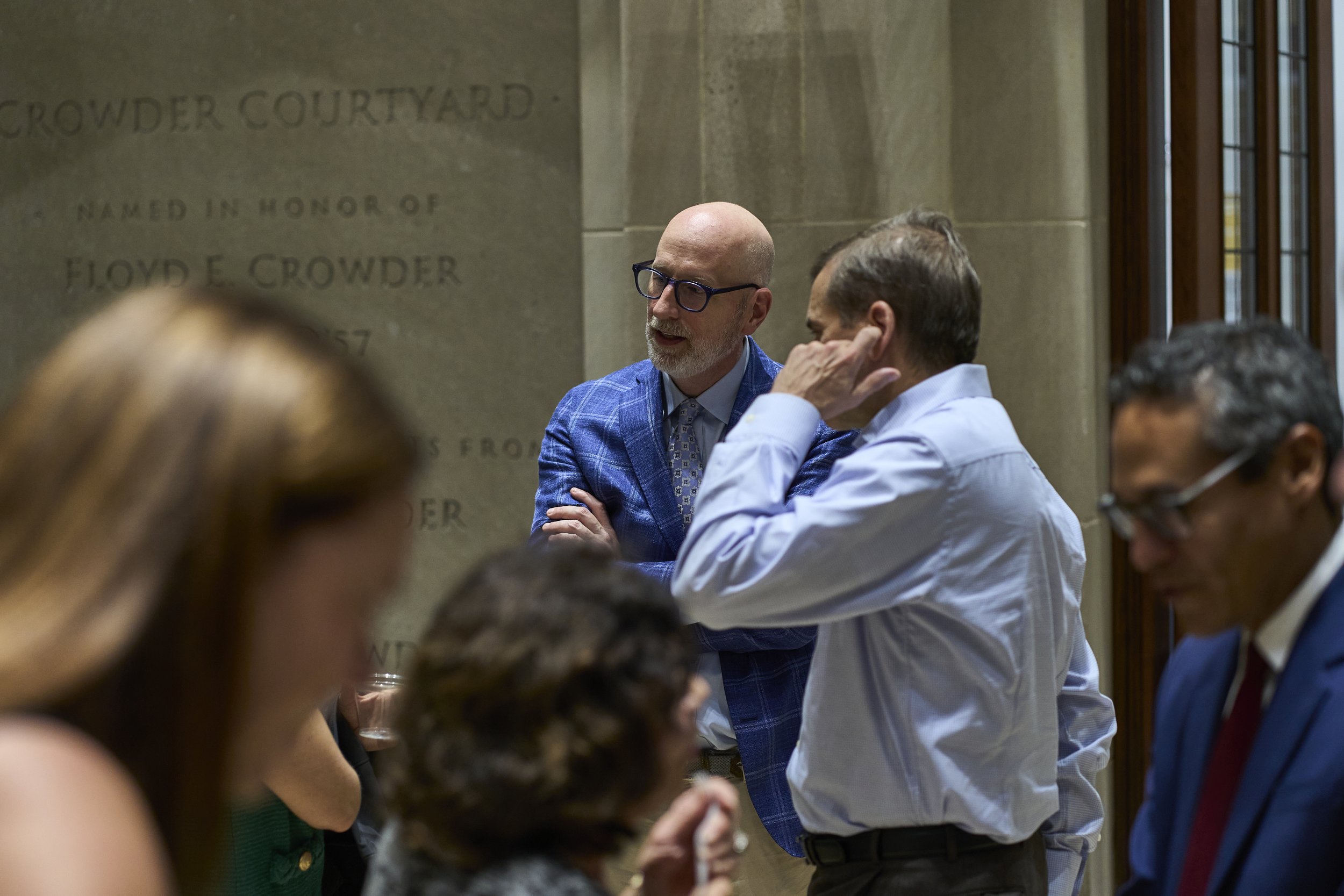 Group of people engaged in conversation in a hallway with a stone wall. A man in a blue plaid blazer and glasses is talking to a man in a white shirt. Other individuals are partially visible.