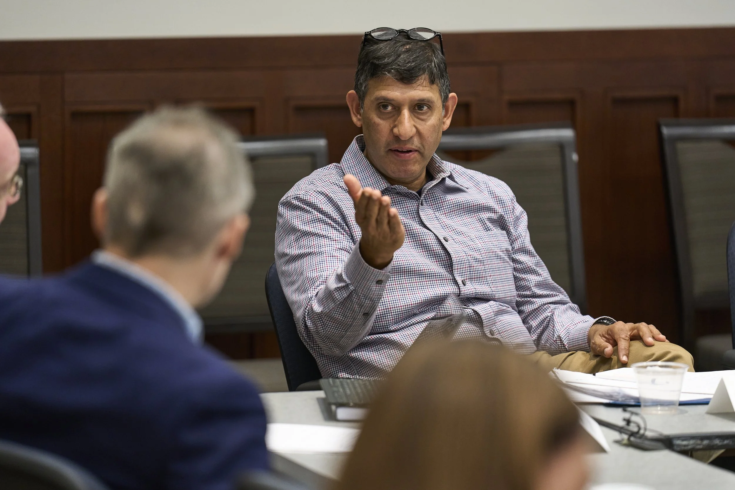 A man in a blue shirt gesturing with his hand in a discussion, sitting at a table in a meeting room.