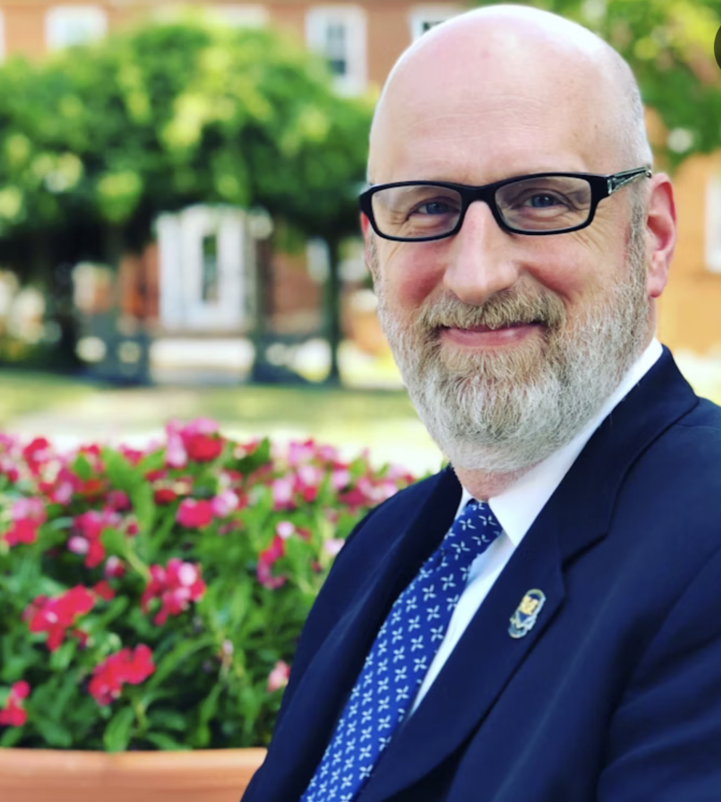 A middle-aged man with a bald head, glasses, and a beard, smiling outdoors in front of a flower bed and a house with trees in the background.