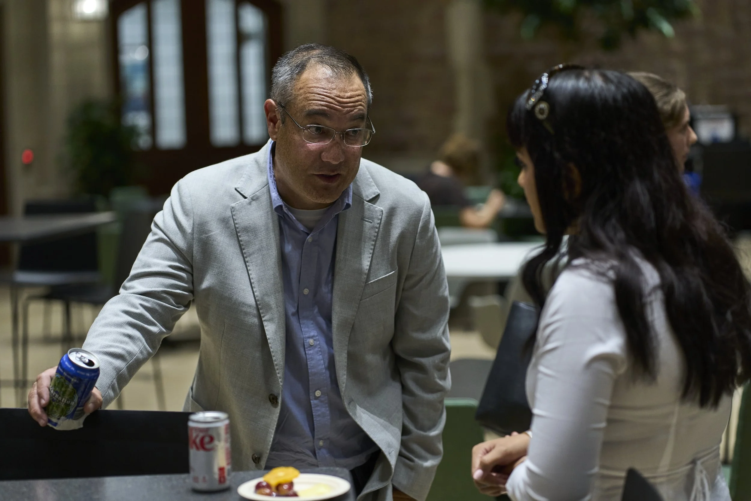 Two people engaged in conversation in a casual indoor setting. One man with glasses in a light gray blazer leaning on a table, holding a can of soda. A woman with dark hair in a white shirt facing him, holding a black folder. There are cans of soda and a dessert on the table.