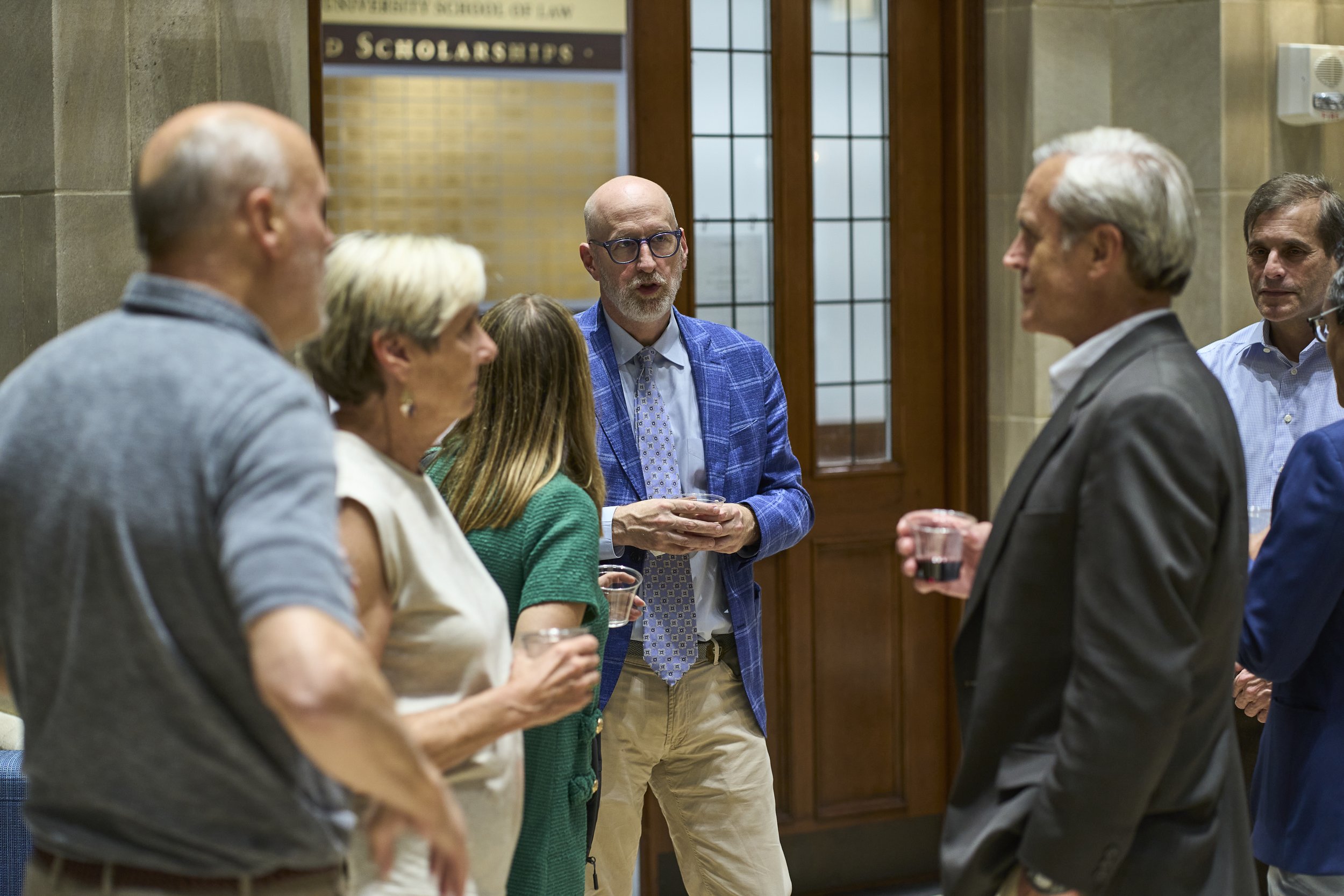 A group of six people, four men and two women, engaged in conversation at a formal indoor event, holding drinks, with wooden doors and a sign about scholarships in the background.