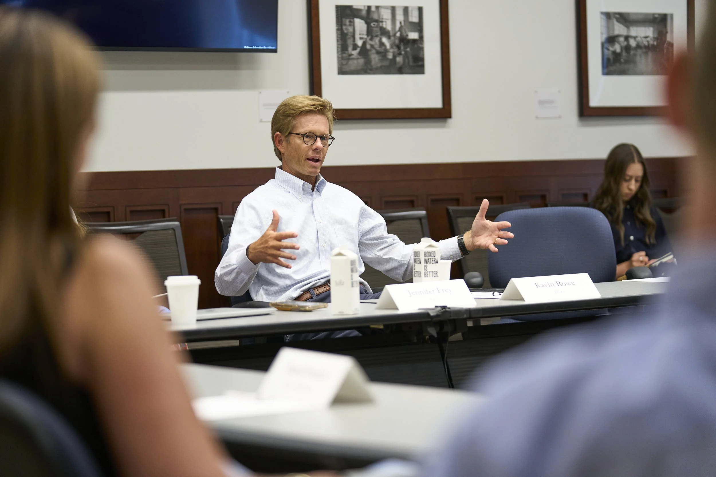 A man with glasses, wearing a white button-up shirt, sitting at a conference table in a meeting room, speaking and gesturing with his hands while seated during a discussion.