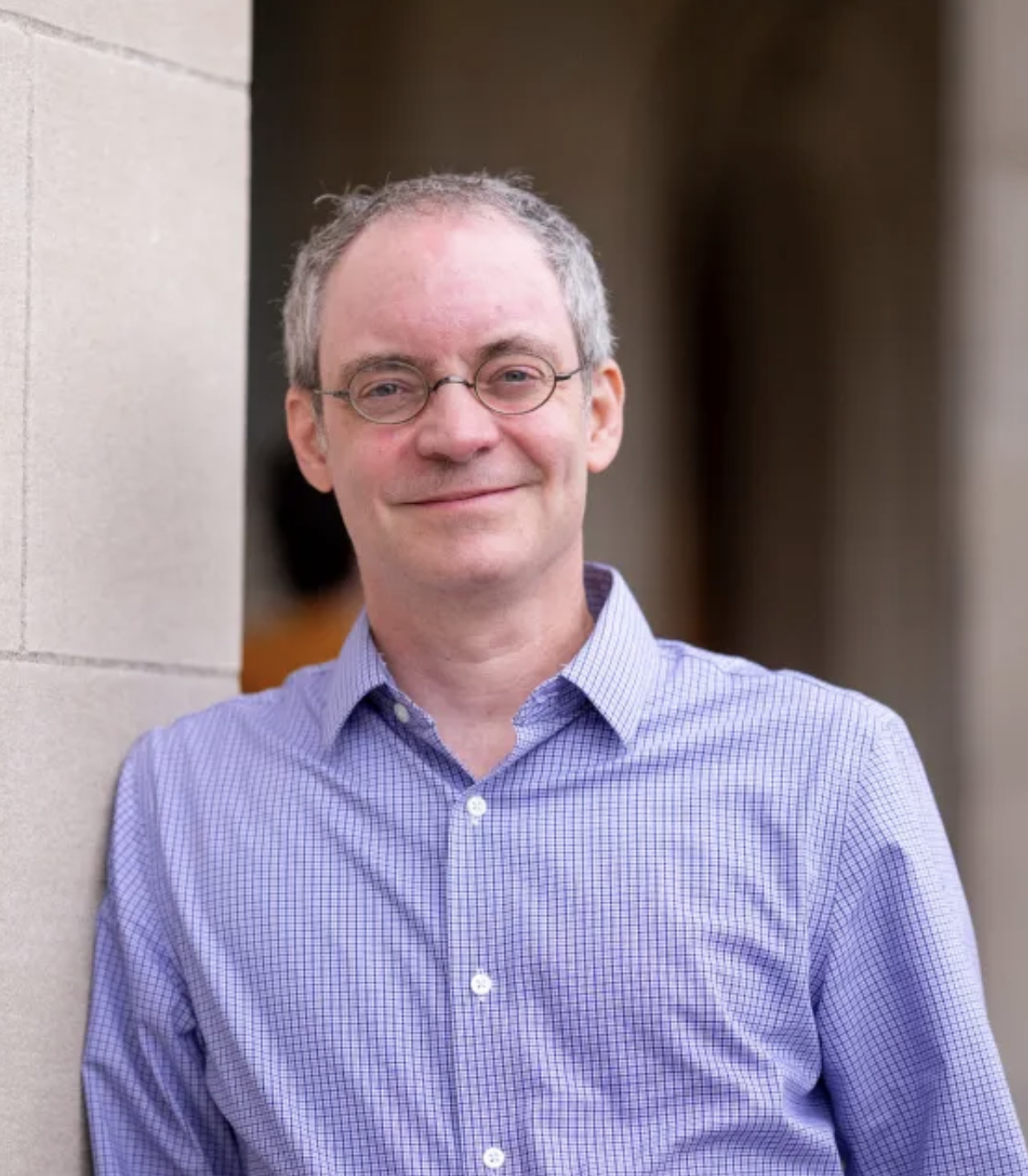 A middle-aged man with glasses and gray hair, wearing a light purple checkered shirt, leans against a wall and smiles at the camera.