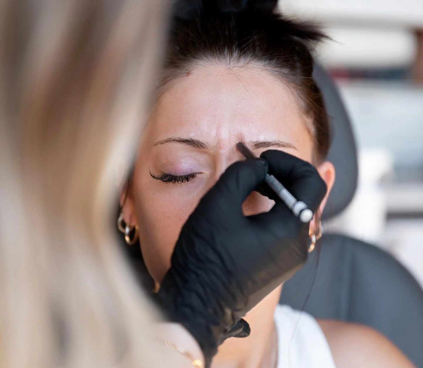 A woman receiving a cosmetic tattoo procedure on her eyebrows from a technician wearing black gloves.