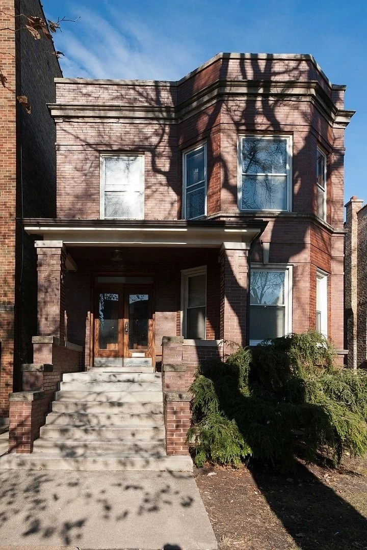 A brick house with a front porch, wooden front door, and large windows. Shadow of tree branches is cast across the house and steps leading up to the porch.