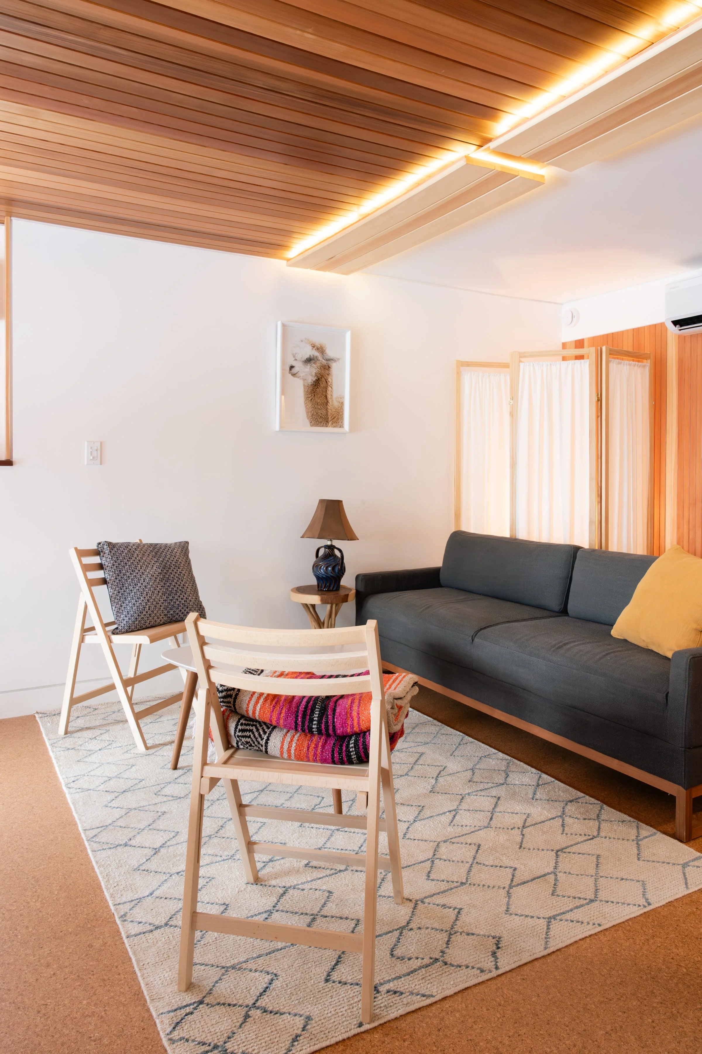 Living room with a dark gray sofa, wooden accent wall, white wall with llama picture, wooden ceiling with recessed lighting, patterned rug, two white chairs with colorful blankets, a side table with a puppet lamp, and a window with light curtains.