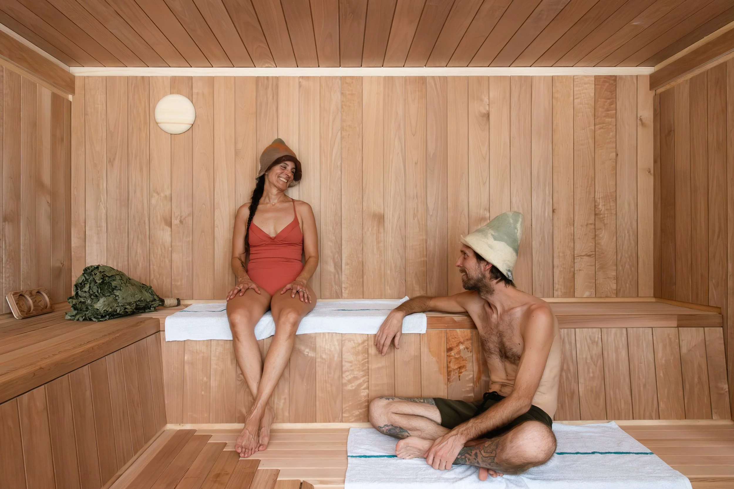 A woman and a man inside a wooden sauna, sitting on benches. The woman is wearing a red swimsuit and a beige sauna hat, smiling and looking at the man. The man is shirtless, wearing black shorts and a beige sauna hat, sitting on the floor on a towel, looking back at the woman and smiling.