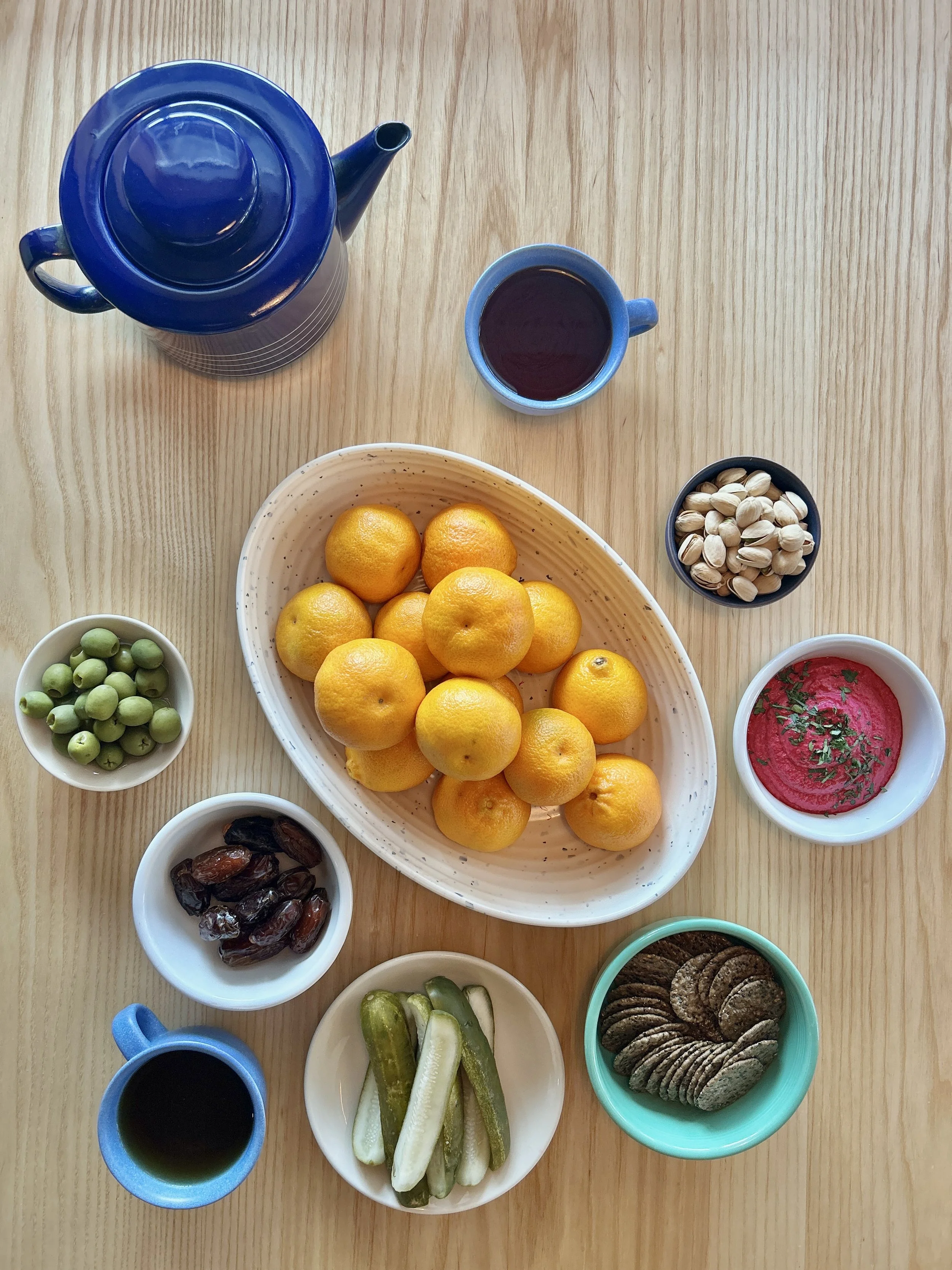 A top-down view of a wooden table with a blue teapot, a mug of dark tea, and a variety of small bowls containing nuts, seeds, pickles, dates, a pink dip with herbs, and cookies surrounding a large oval white bowl of yellow citrus fruits.