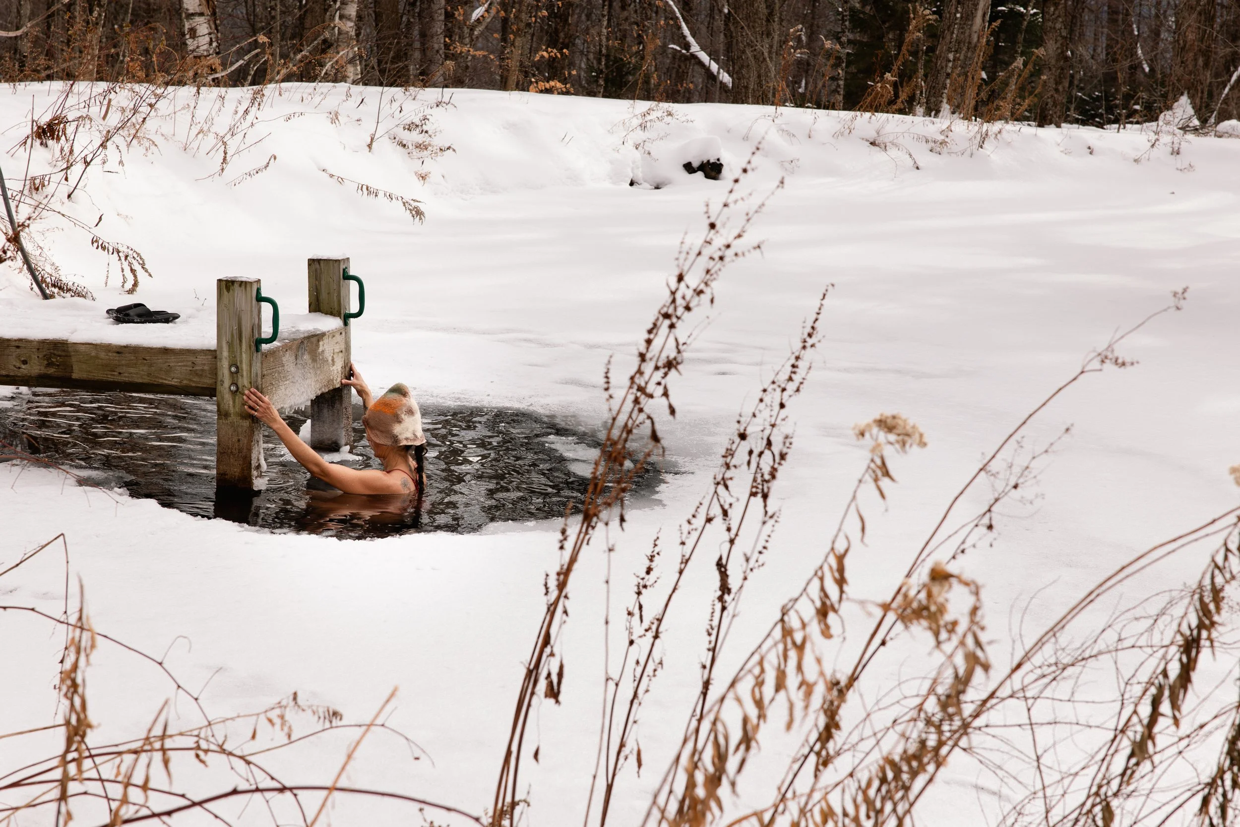 Person with a hat swimming in an ice-covered hole in a frozen lake in winter, holding onto a wooden dock on a snowy landscape.