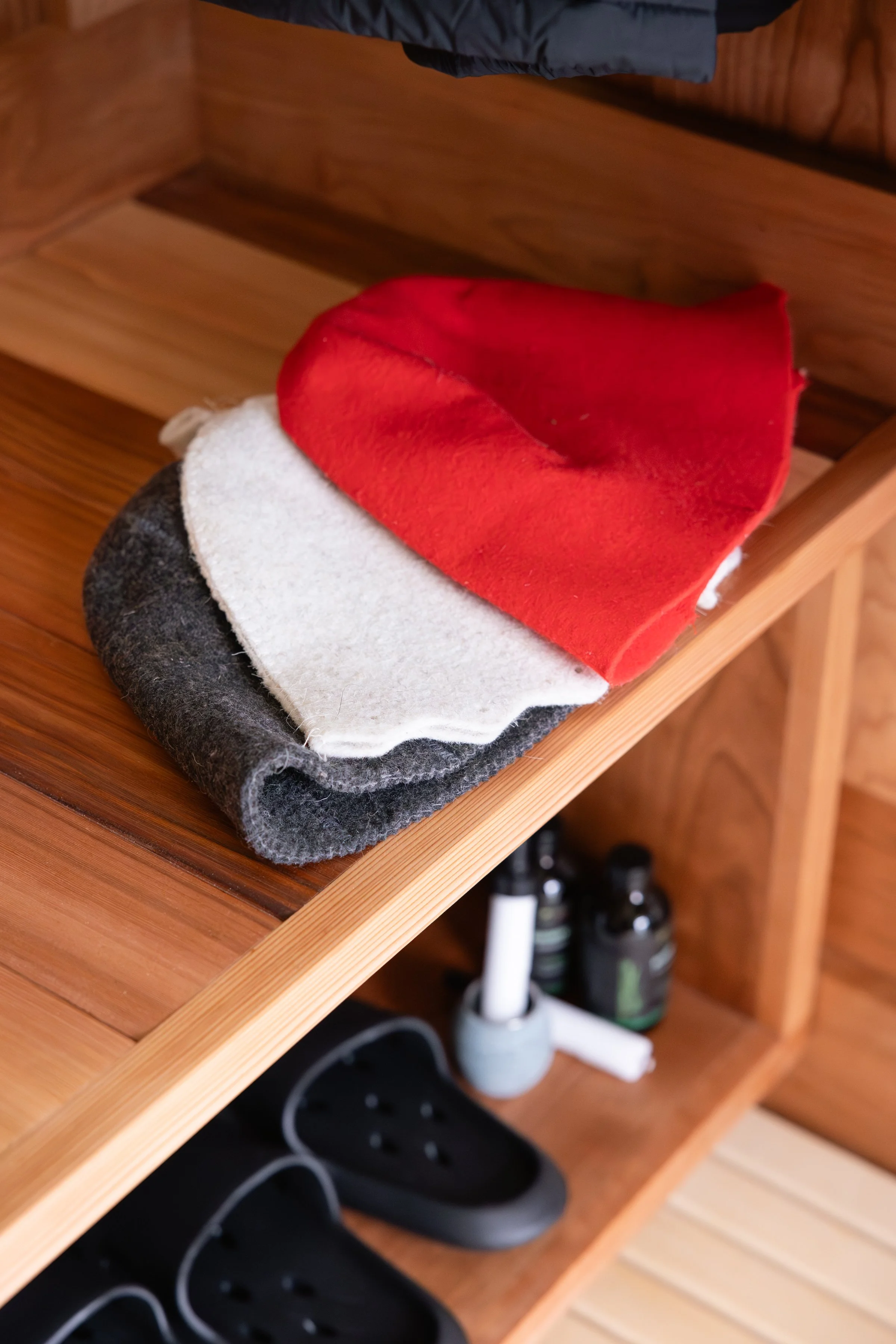 A wooden shelf containing three cleaning or polishing cloths in red, white, and black, with black slippers and bottles of cleaning supplies below.