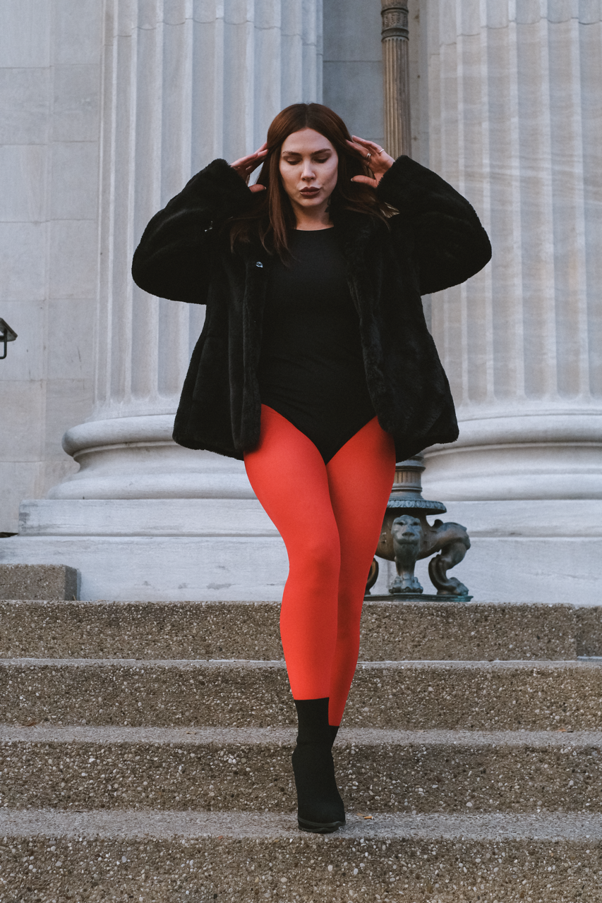 A woman with brown hair in a black fur coat and black top, wearing bright red tights and black ankle boots, walking down stone stairs in front of a historic building with large columns.