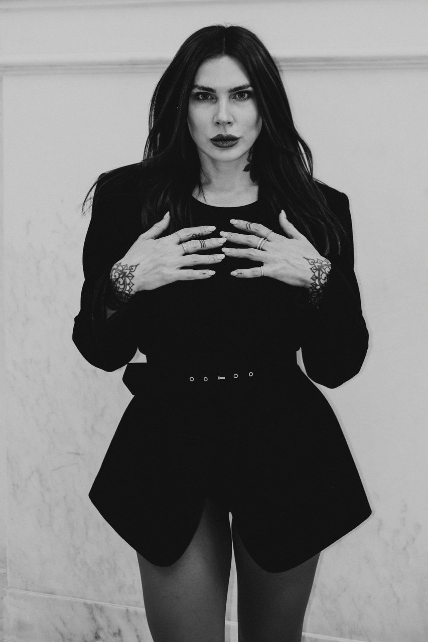 A woman with dark hair and tattoos on her hands, wearing a black outfit, standing indoors against a plain wall, looking directly at the camera.