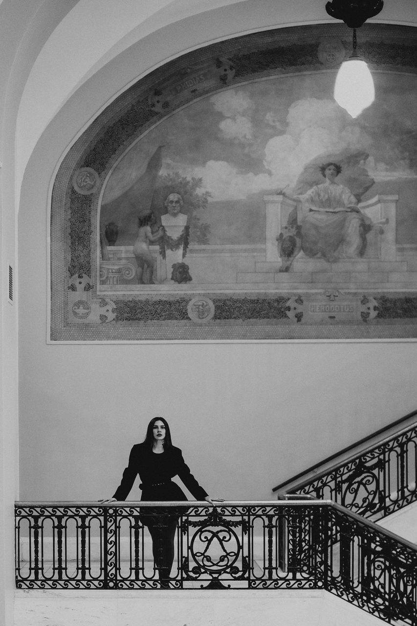 A woman with dark hair, dressed in black, standing on a staircase with an ornate railing in an interior space, with a large fresco painting on the wall above her.